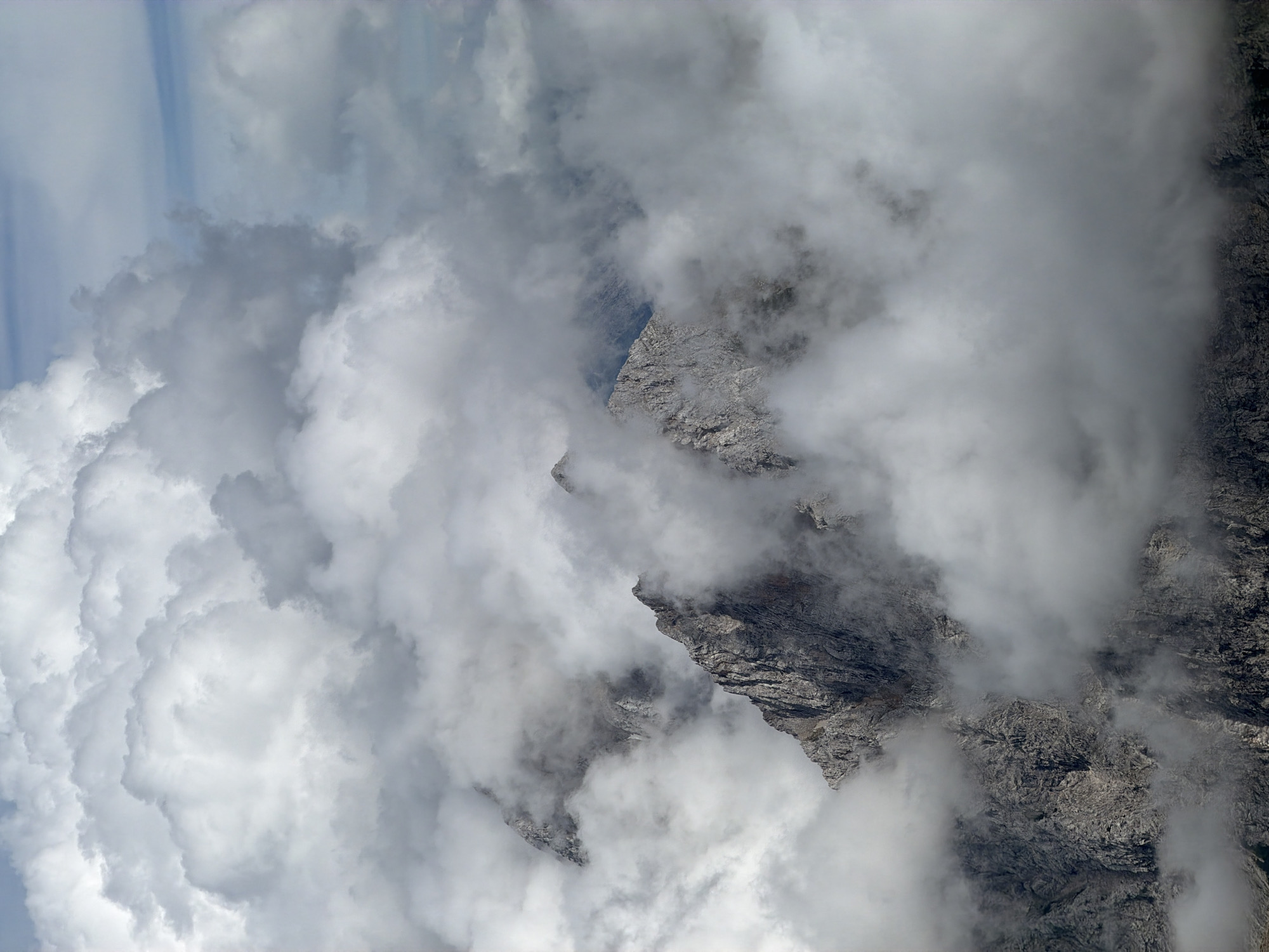 This image captures a dramatic view of mountain peaks emerging from a thick blanket of clouds, suggesting a high-altitude environment. The rugged, rocky texture of the mountains is visible through breaks in the swirling white and grey clouds. The lighting is diffused, indicating an overcast or misty day. The scene evokes a sense of wilderness and the power of nature, with the mountains appearing as solid masses against the ephemeral clouds. The location is likely in the vicinity of Garmisch-Partenkirchen, Germany, a region known for its alpine landscapes. There are no people or specific man-made objects discernible in the image.