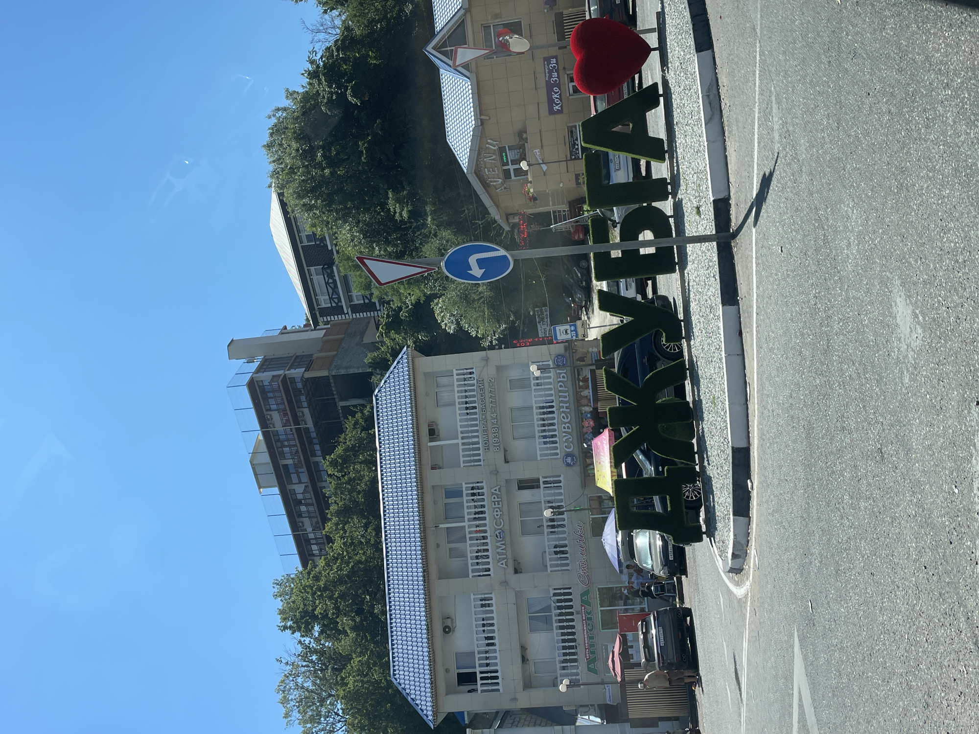 A sunlit, low-angle medium shot captures a street scene in Dzhubga, Russia. Dominating the foreground are large, green, topiary letters spelling "ДЖУБГА," with a large red heart sculpture positioned to the right of the letters. The letters are situated on a gravelly, circular median bordered by a curb. The road surface is asphalt, with a white lane marking.
Behind the letters, a multi-story building with a blue tiled roof stands prominently on the left, featuring white balconies and signs that read "АТМОСФЕРА" and "НОМЕРА-БАССЕЙН". Further to the right, a smaller building with a similar blue roof has a sign that says "AVENU" and "Коко Зи-зи". A street sign with a white triangle and red border is visible above a circular blue sign with a white arrow pointing left.
Several cars are parked or driving on the road. A man in a light-colored shirt and shorts stands near a black SUV on the left side of the frame. In the background, more buildings and dense green trees cover the hillside under a clear blue sky. The overall scene depicts a busy street on a bright day in the Russian coastal town.