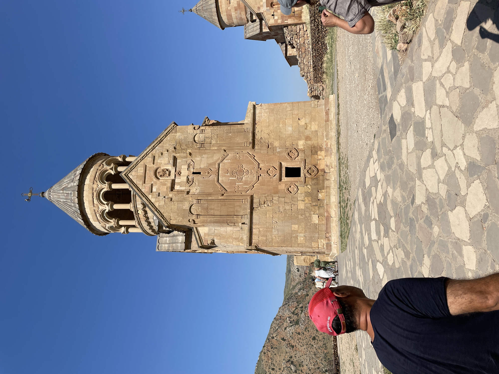 The image shows the exterior of a stone church in Areni, Armenia, under a clear blue sky. A man wearing a red baseball cap with the word "PORSCHE" is in the foreground, facing away from the camera and looking towards the church. He is wearing a dark t-shirt. Behind him, a group of people are walking towards the church, some of whom are tourists. The church is made of light-colored stone and features intricate carvings and a domed roof topped with a cross. Another smaller building, also made of stone, is visible to the right of the main church. The ground in front of the church is paved with irregular stone slabs. In the background, a rocky, mountainous landscape is visible.