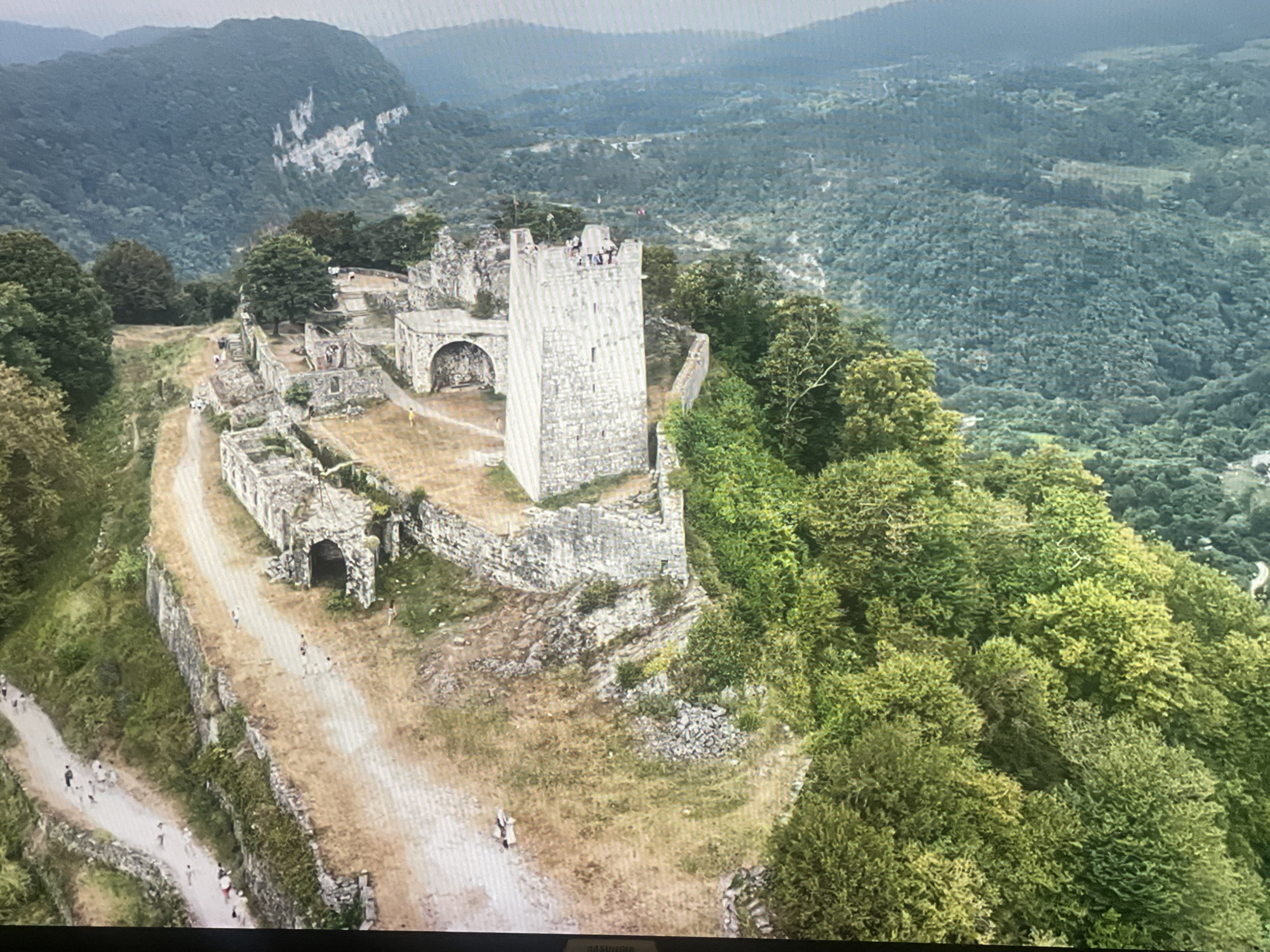 The image shows the ruins of a stone fortress situated on a hilltop, surrounded by lush green forests and mountainous terrain. The fortress features a prominent square tower and several ruined walls and archways. Several people, appearing as small figures from this aerial perspective, are visible walking along pathways within the fortress complex and on the surrounding grounds. The overall environment suggests a historical site in a natural, somewhat remote setting. This is likely the Anacopia Fortress, located in New Athos (New Afon), a resort town in Abkhazia, Georgia, near the city of Sukhumi.