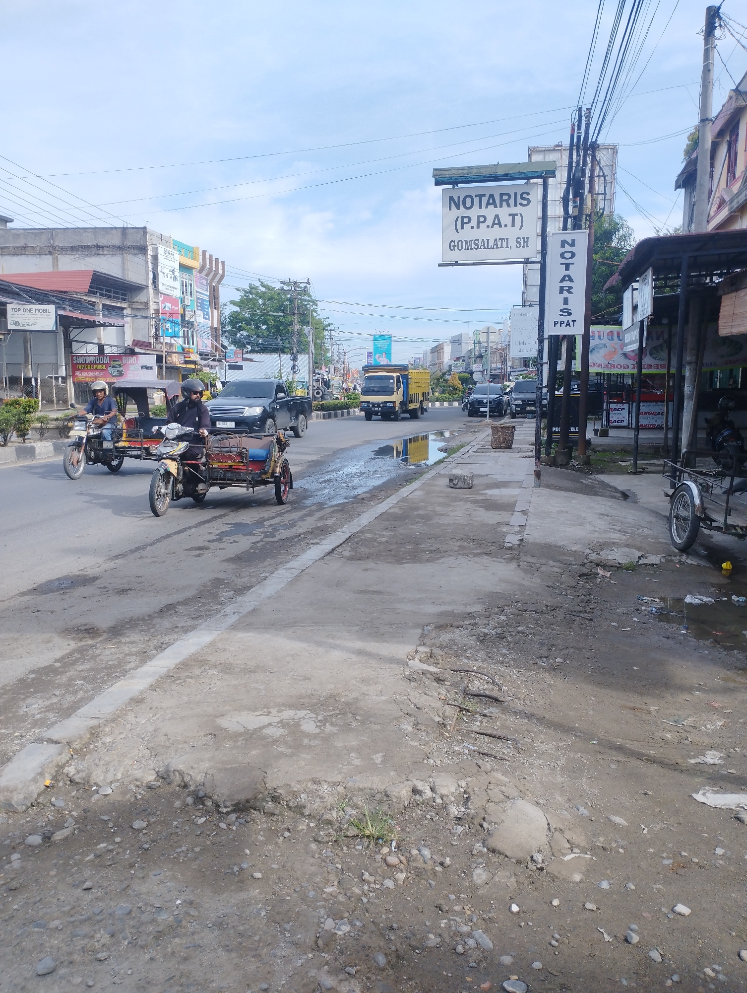 The image depicts a street scene in Lhokseumawe, Indonesia. A yellow truck and several other vehicles, including pickup trucks, are visible on the road. Two motorcycle rickshaws are moving on the road. On the right side of the frame, a sign reads "NOTARIS (P.P.A.T) GOMSALATI, SH." Another narrower vertical sign also indicates "NOTARIS PPAT." Adjacent to the sign, there's a building or shop with advertisements and information posted, along with a parked three-wheeled vehicle. The sidewalk is cracked and shows signs of disrepair, with visible stones and debris. Overhead, electrical wires crisscross the sky. Buildings and storefronts line the street in the background. The sky is partly cloudy.