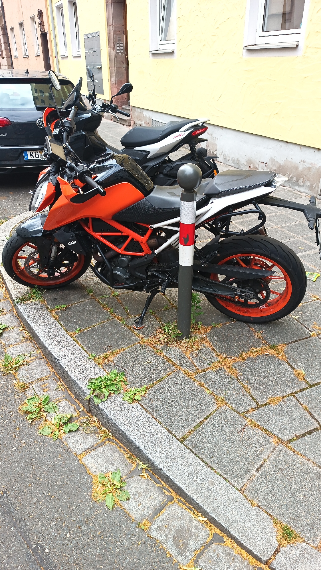 The image shows two motorcycles parked on a cobblestone sidewalk next to a street in Nuremberg, Germany. The foreground motorcycle is an orange KTM Duke, partially obscuring a white and black scooter parked behind it. A black Volkswagen Golf is visible on the left edge of the frame. The buildings in the background are painted in yellow and feature red-brown accents around windows and doors. A bollard with red and white stripes stands between the two motorcycles. The ground is paved with gray cobblestones and bordered by a granite curb, with weeds growing in the cracks.