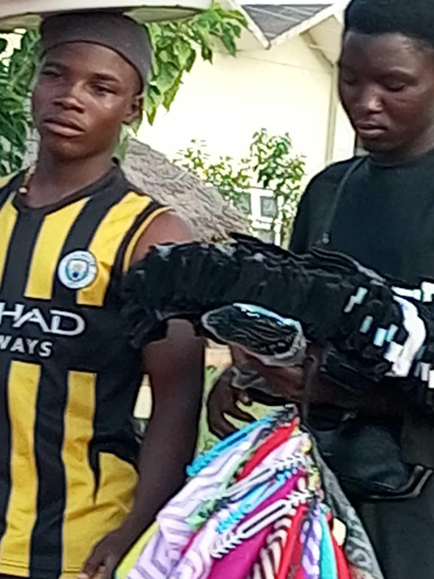 The image shows two young Black men in Gombe, Nigeria. The man on the left is wearing a black and yellow striped jersey with the Manchester City Football Club logo and the word "HAD" visible, along with a dark cap. He appears to be holding a bundle of dark material, possibly fabric or clothing, with white accents. The man on the right is wearing a black t-shirt and appears to be holding a different bundle of colorful textiles, possibly scarves or wraps, with white plastic fasteners. The background suggests an outdoor setting, possibly a market, with foliage and a building visible. The overall scene depicts a commercial transaction or display of goods.