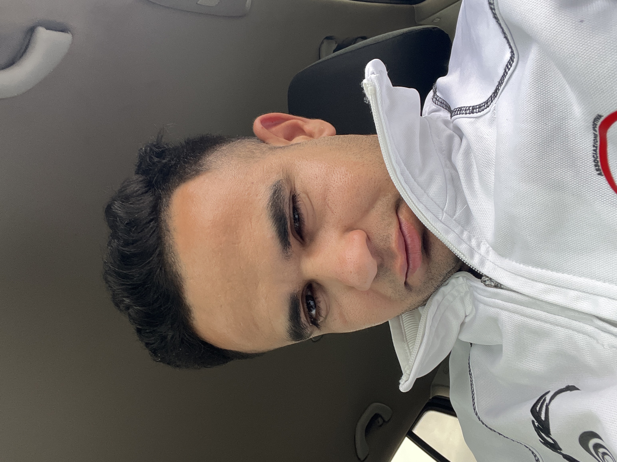 A young man is depicted in a close-up, selfie-style shot taken inside a vehicle. He has dark hair, dark eyes, and a light tan complexion with short stubble around his jawline. His gaze is directed towards the camera with a calm, slightly softened expression.
He is wearing a white zippered jacket or sweatshirt with a textured fabric. Black stitching or piping is visible along the seams of the garment, particularly on the shoulder area. Partially visible on the right side of his chest is text that reads "ASSOCIAZIONE PRO..." (likely short for "Associazione Sportiva" or similar, suggesting an Italian sports association), alongside a black stylized design that could be a logo or part of a larger graphic.
The setting is the interior of a car or similar vehicle, indicated by the dark grey headliner and an overhead grab handle visible in the background. Parts of dark car seats are also visible. The lighting suggests it is daytime, with some light entering from a window. The man appears to be seated, possibly as a passenger or driver at rest. The scene is set in Martil, Morocco.