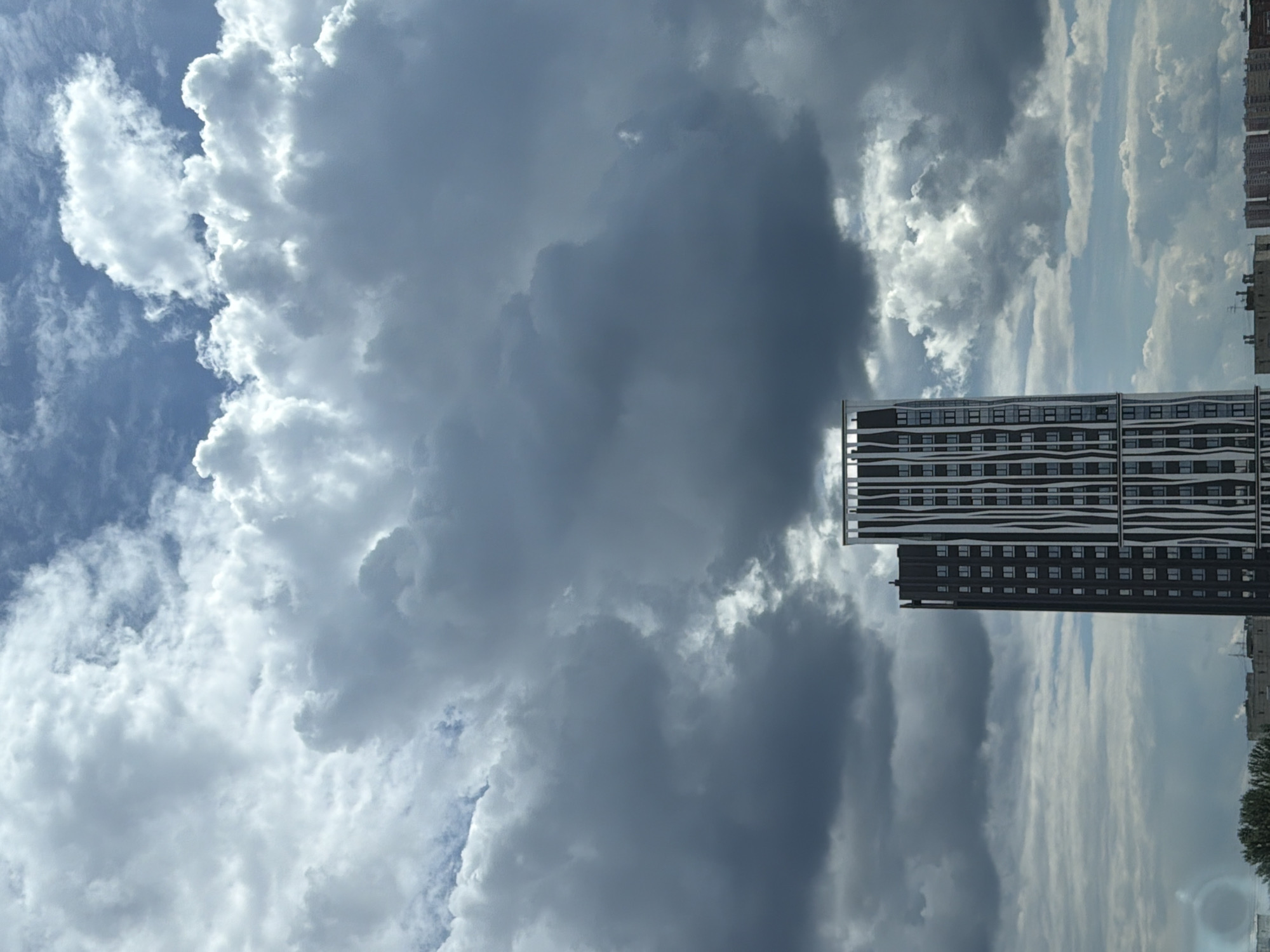This is a vertical, eye-level medium shot of a city skyline with a dramatic sky. The primary subjects are two modern buildings with dark facades and a complex pattern of windows. The building in the foreground is taller and appears to be a residential complex, featuring a striking wavy pattern in its facade. The building behind it is shorter and darker. The setting is urban, with hints of a cityscape on the right side of the frame, including what appears to be a tree and the tops of other buildings.
The sky is the dominant element, filled with a mix of dark, heavy storm clouds and brighter, wispy clouds illuminated by sunlight. The contrast between the dark clouds and the bright, sunlit areas creates a sense of dynamic weather, possibly a storm approaching or passing. The overall atmosphere is dramatic and a bit ominous due to the dark clouds, yet the bright areas suggest moments of light breaking through.
There is no visible activity or event taking place; it's a still scene focused on the architecture and the sky. The time of day is suggested by the quality of light, likely daytime with overcast conditions and sun breaking through. The location is identified as Perm, Russia. No text is visible in the image.