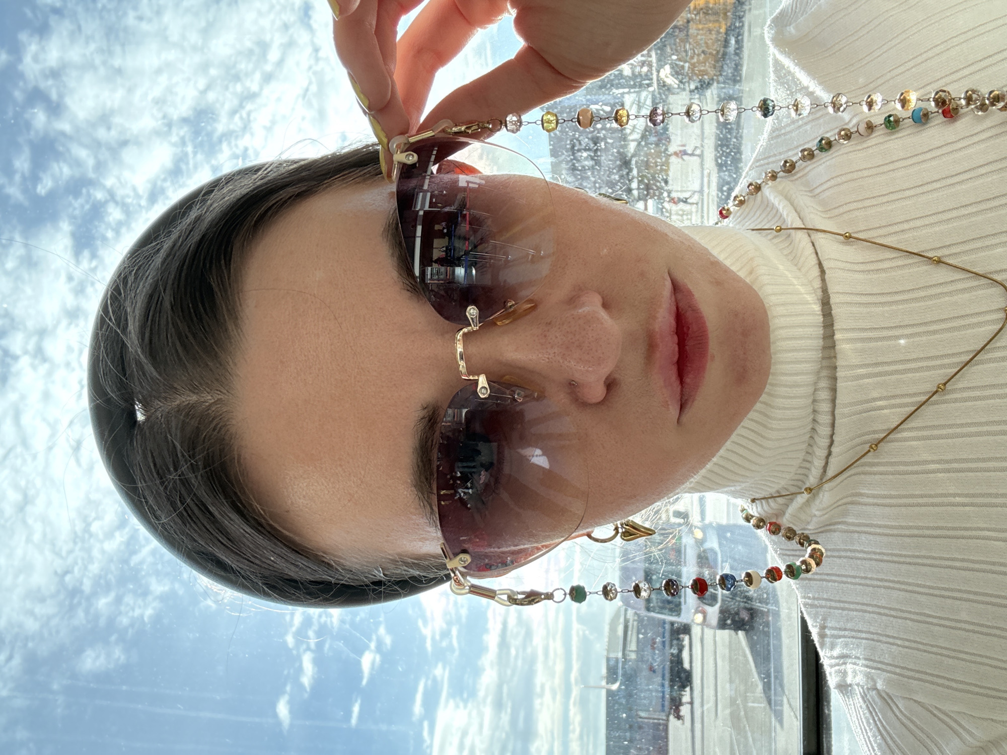 A woman is holding up a pair of rimless, light-tinted sunglasses with a gold frame and a beaded chain attached in front of a window. The woman has dark hair and is wearing a white turtleneck sweater. The sunglasses reflect what appears to be the interior of a building. Through the window, the sky with clouds can be seen. There are blurred buildings and vehicles outside the window. Based on visual analysis, it is impossible to definitively identify the location as the city of Khimki, Russia.