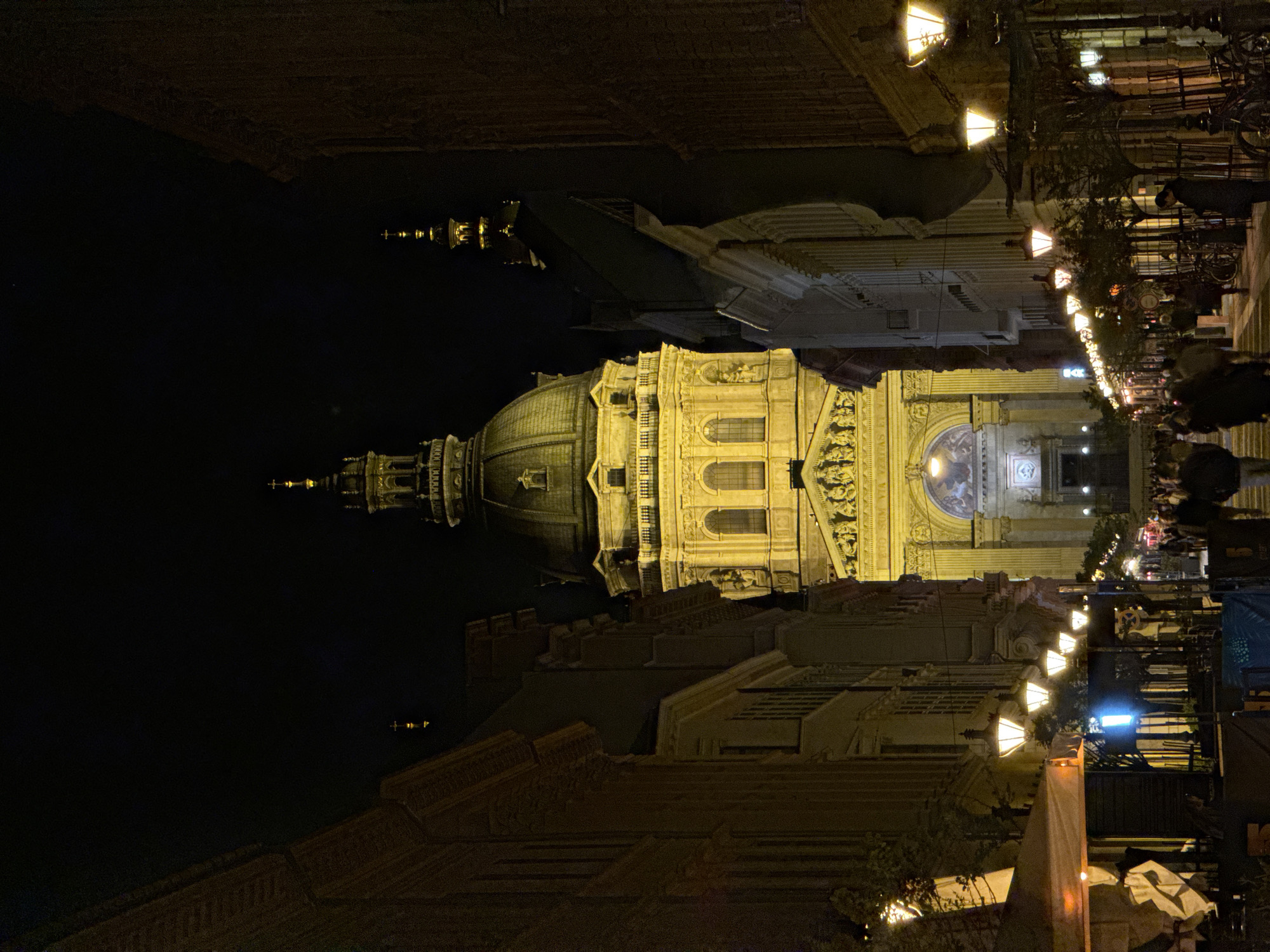 Nighttime view of St. Stephen's Basilica in Budapest, Hungary. The basilica is brightly lit, contrasting with the dark sky. Street lamps line the walkway in front of the basilica, illuminating the area where pedestrians are walking. The architecture surrounding the basilica is also visible. The image is taken from a ground-level perspective.