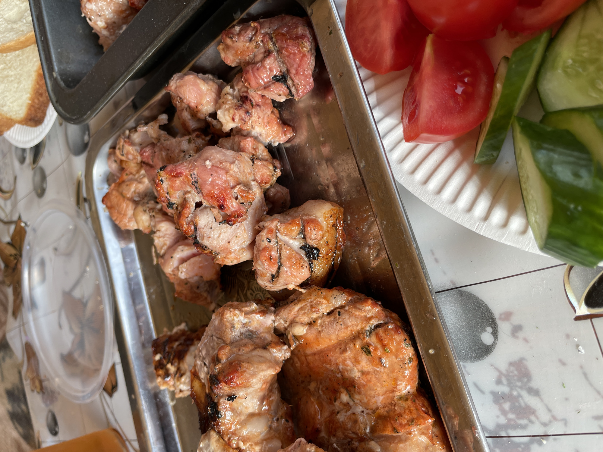 A close-up, overhead shot captures a meal laid out on a table, likely outdoors given the lighting. In the foreground, a white paper plate holds sliced tomatoes and cucumbers, suggesting a fresh salad. To the left, a metal tray is filled with pieces of cooked meat, appearing to be grilled or roasted, with some pieces showing char marks. A transparent plastic plate with remnants of food is visible in the background on the left. To the right, a dark metal baking pan contains more meat, possibly raw or precooked, and beside it, two slices of white bread sit on a small white plate. The table surface is covered with a patterned tablecloth. The overall scene suggests a casual gathering or meal preparation. This image does not contain any information that would allow it to be identified as being located in Novokuznetsk, Russia.
