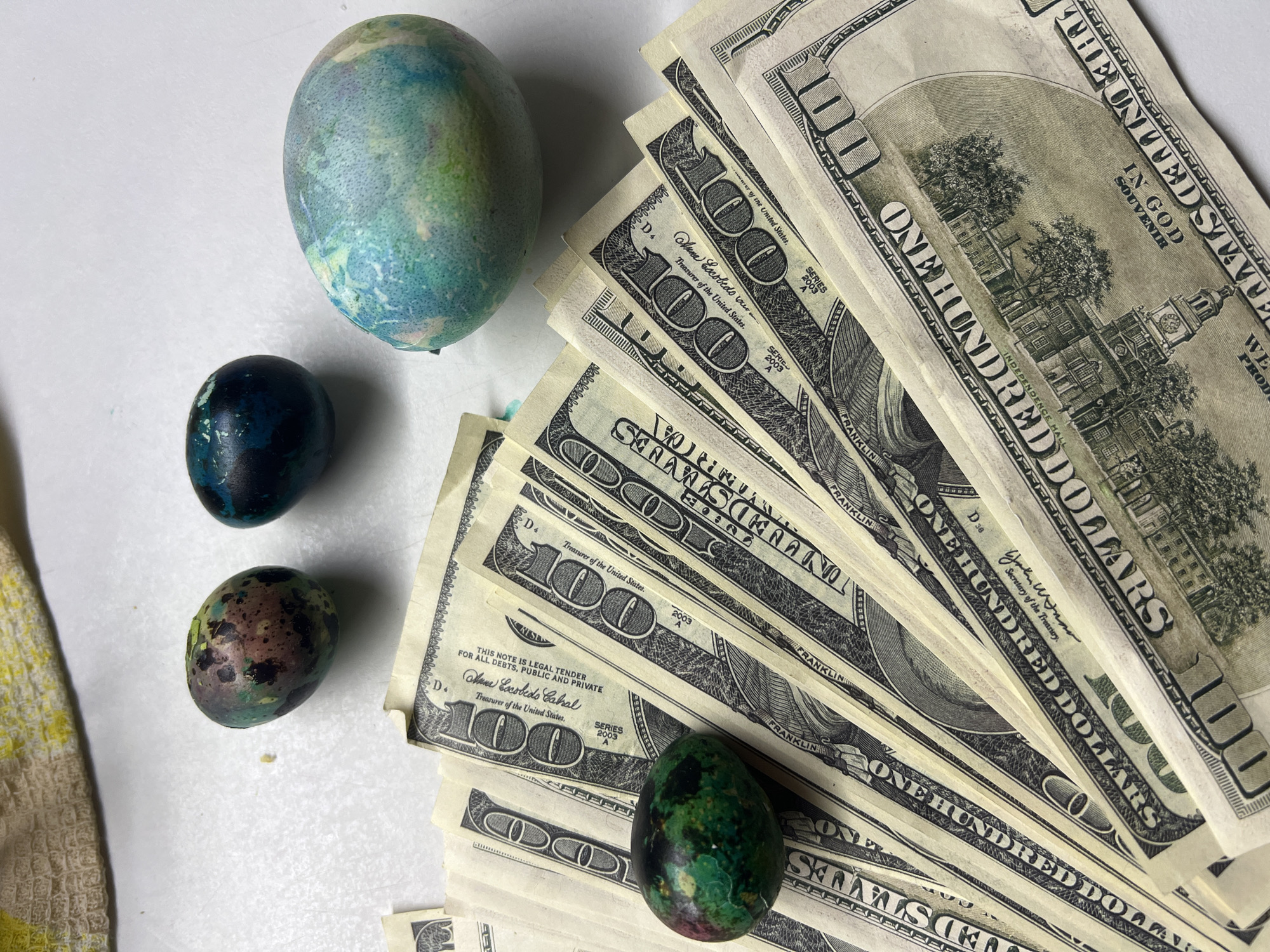 The image shows a collection of objects on a white surface. There are four painted Easter eggs, with varying color schemes of blue, green, and brown. Adjacent to the eggs is a stack of United States one hundred dollar bills. A small portion of a textured yellow and white cloth is visible in the lower left corner. The image contains no information about the city of Noviy, Russia.