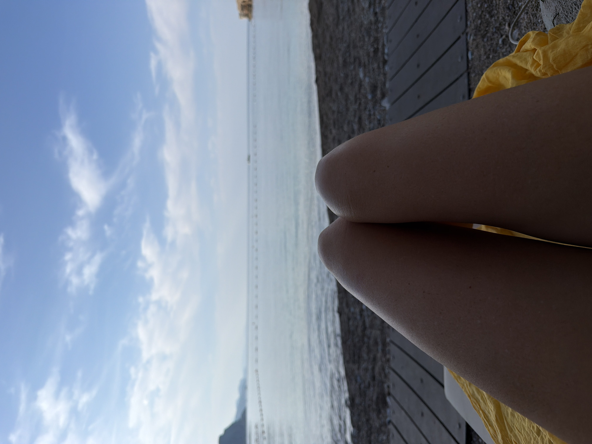 The image captures a scene from a beach, likely in Budva, Montenegro, based on the landscape visible in the background. The perspective is that of someone lying down, with their legs in the foreground. The legs are resting on what appears to be a wooden deck or sunbed, with a yellow piece of fabric or clothing partially visible. Beyond the legs and deck, the image shows the sea, with buoys visible in a line across the water. The shoreline is visible on the right side, leading to a mountainous headland in the distance on the left. Above, the sky is light blue with scattered white clouds.