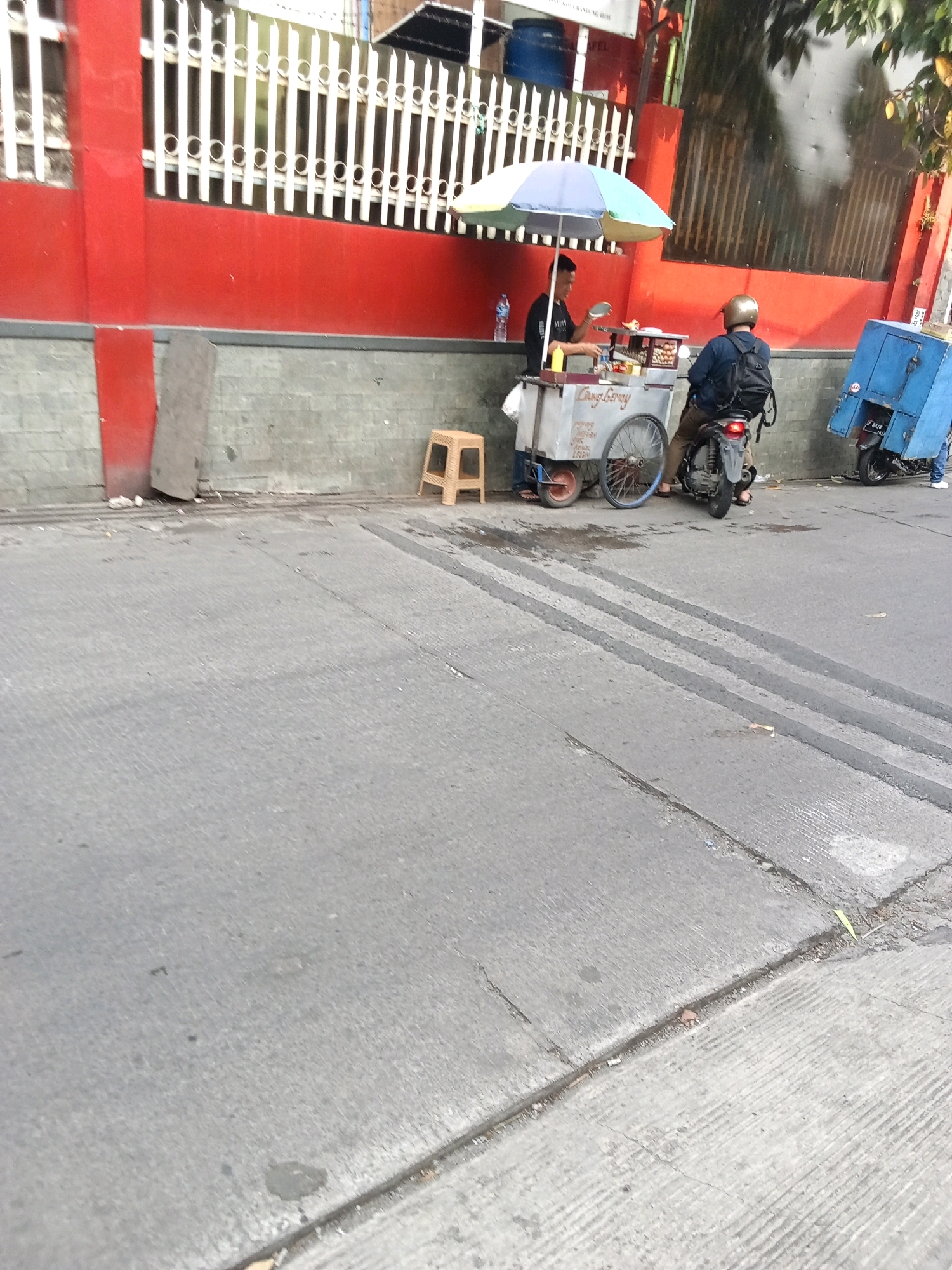 The image shows a street scene, likely in Bandung City, Indonesia, with a food vendor's cart and two individuals. A man is standing behind the cart, preparing food, while another person sits on a motorcycle. The food cart is equipped with an umbrella for shade. A blue box is attached to a motorcycle parked further down the street. The environment appears to be an urban setting with a red-painted wall and a metal fence in the background. The foreground consists of a concrete road with visible tire tracks. The scene suggests a typical street-side food vending operation.