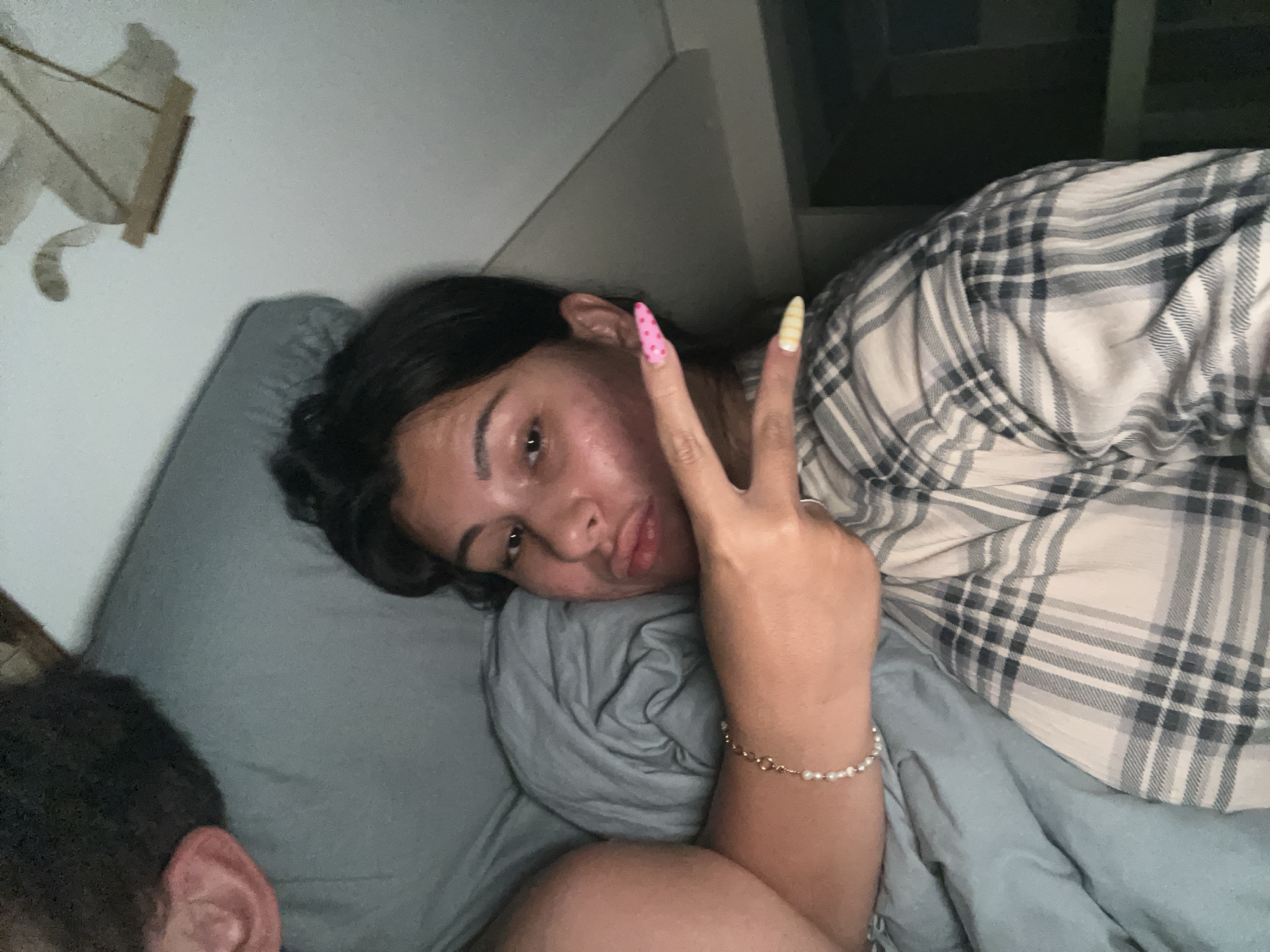 A close-up, dimly lit indoor photograph, taken in Kiryat Bialik, Israel, features a young adult female lying on her left side, her head resting on a grey pillow. She has dark hair, a medium complexion, and is looking directly at the camera with a slight pout. Her right arm is raised, forming a peace sign with her index and middle fingers. The nail on her index finger is pink with white polka dots, and the nail on her middle finger is yellow with white stripes. A delicate pearl bracelet adorns her right wrist. She is wearing a plaid shirt with light and dark grey checks. Her lower body and the surrounding bedding appear to be covered by a solid blue-grey duvet or blanket. In the bottom left corner, the top of a second person's head and a right ear are partially visible, indicating another individual lying nearby. In the upper left background, an out-of-focus, light-colored cutout resembling an animal is affixed to a white wall. The upper right background is dark and indistinct, possibly a wall or furniture.