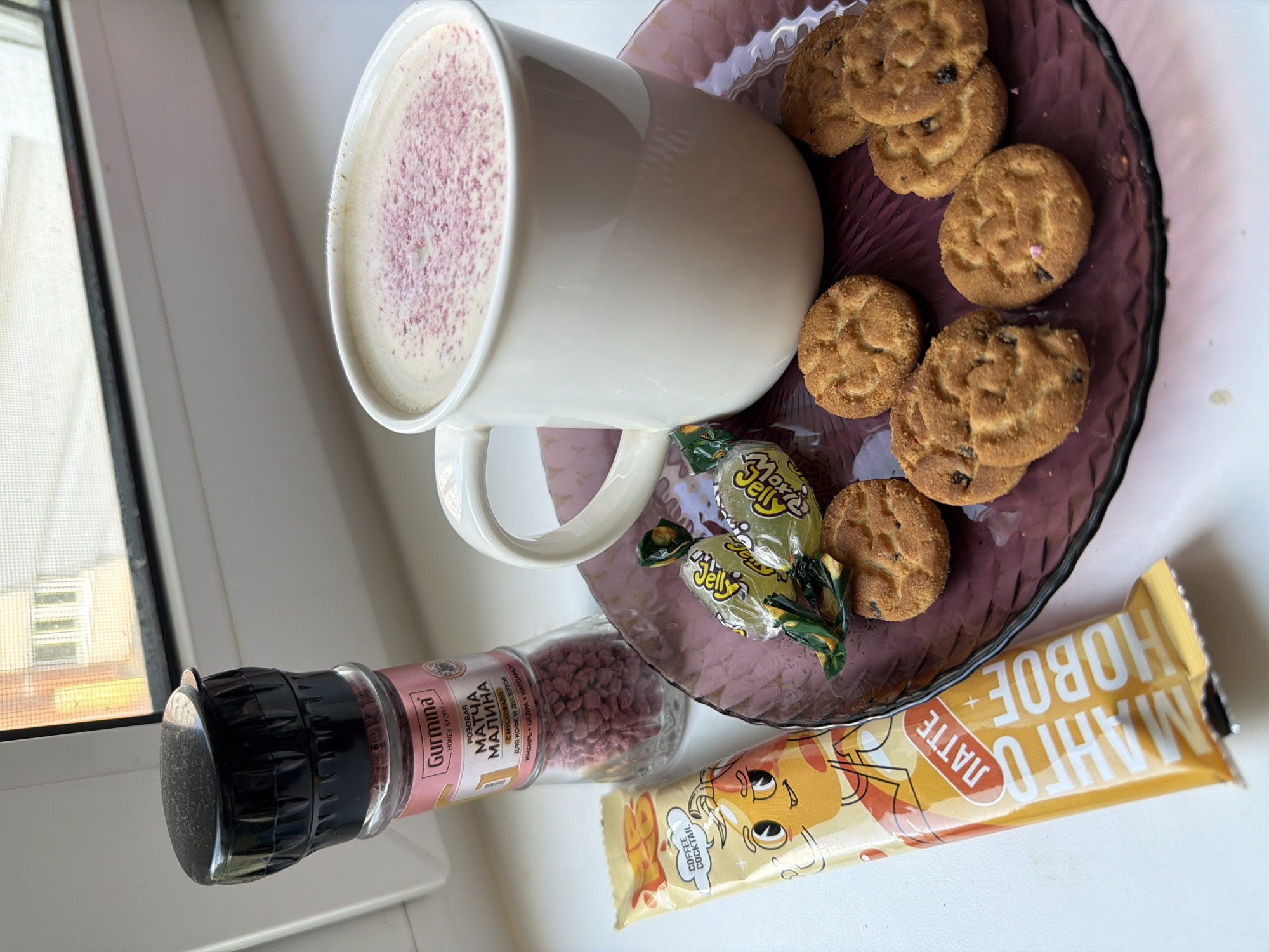 The image depicts a close-up, overhead view of a collection of food items arranged on a windowsill, possibly in a residential setting. In the foreground, on the left, is a yellow, foil-wrapped snack bar with Russian text that translates to "Mango Latte." To the right of the bar is a clear glass shaker filled with pink granular material, labeled in Russian as "Gurmina Honey Story Rose Matcha Raspberry." This shaker is positioned behind the snack bar.
Dominating the right side of the frame is a white ceramic mug filled with a frothy beverage, possibly a latte or cappuccino, lightly dusted with pink powder. The mug rests on a translucent, dark purple, patterned glass plate. Arranged on the plate are several round, golden-brown cookies, some with visible dark inclusions that appear to be raisins or chocolate chips. Two individually wrapped, yellow candies with green accents are placed on the plate near the mug.
The background shows a blurred view of a window with a white frame, through which the upper floors of a building with windows are visible. The overall lighting suggests an indoor environment, possibly during daylight hours. The specific location in the city of Chaikovsky, Russia, cannot be determined from the visual information provided in the image itself.