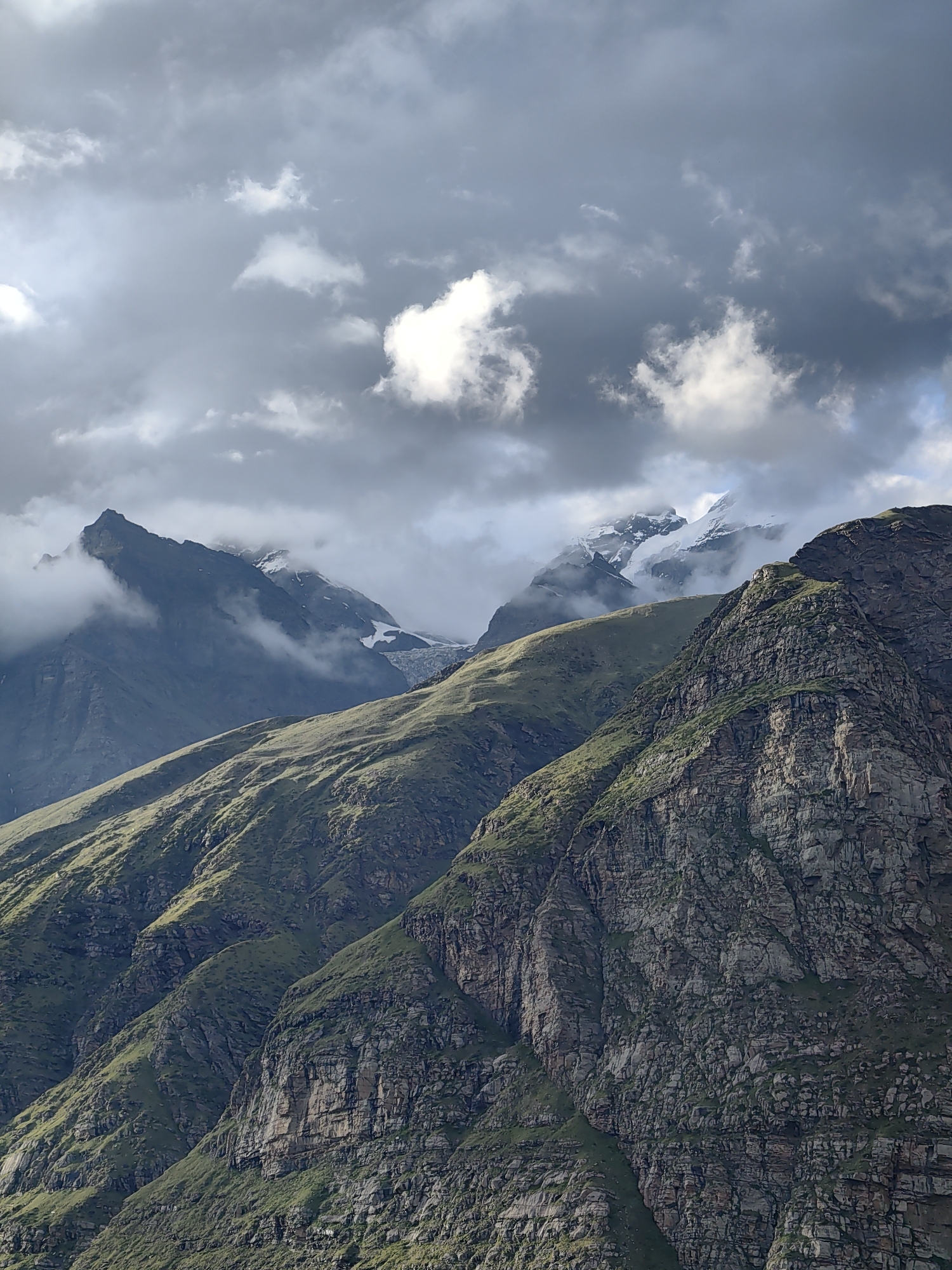 This is a vertical, medium shot of a mountainous landscape in Gramphu, India, on a cloudy day. The foreground is dominated by a steep, rocky mountainside covered in sparse green vegetation, characterized by its textured surface and visible layers. In the mid-ground, a gentler, grass-covered slope ascends, its contours highlighted by the play of light and shadow. Further back, imposing, darker peaks rise, partially shrouded in mist and clouds, with some snow-capped summits visible in the distance. The sky is filled with dramatic, gray clouds, with patches of brighter sky and sunlight breaking through, casting a dynamic light on the scene. The image captures a rugged, natural environment with a sense of immense scale.