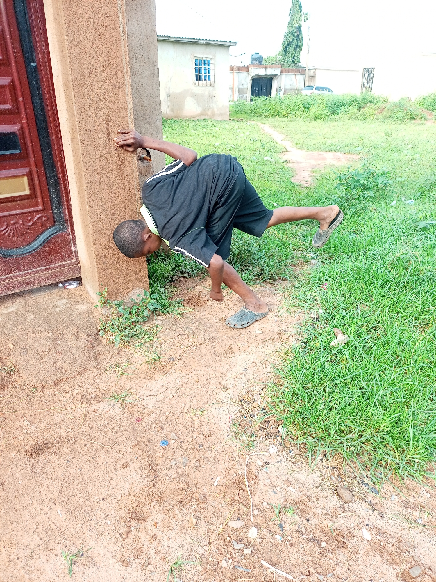 In Tempure, Nigeria, a young boy is engaged in the game of "hide and seek" against a rough-textured, sandy-colored wall. He is leaning forward, with his head pressed against the wall, and his body is angled such that his legs are extended backward. The boy is wearing a black t-shirt with white stripes on the sleeves and black shorts. He has on a pair of dark, open-toed sandals. The ground is a mix of sandy soil and patches of green grass. In the background, a building with a white facade and a blue-paned window is visible, along with other structures and dense vegetation. A tall, slender tree stands in the distance. The overall scene suggests an outdoor setting in a residential area, with a clear indication of a game being played.