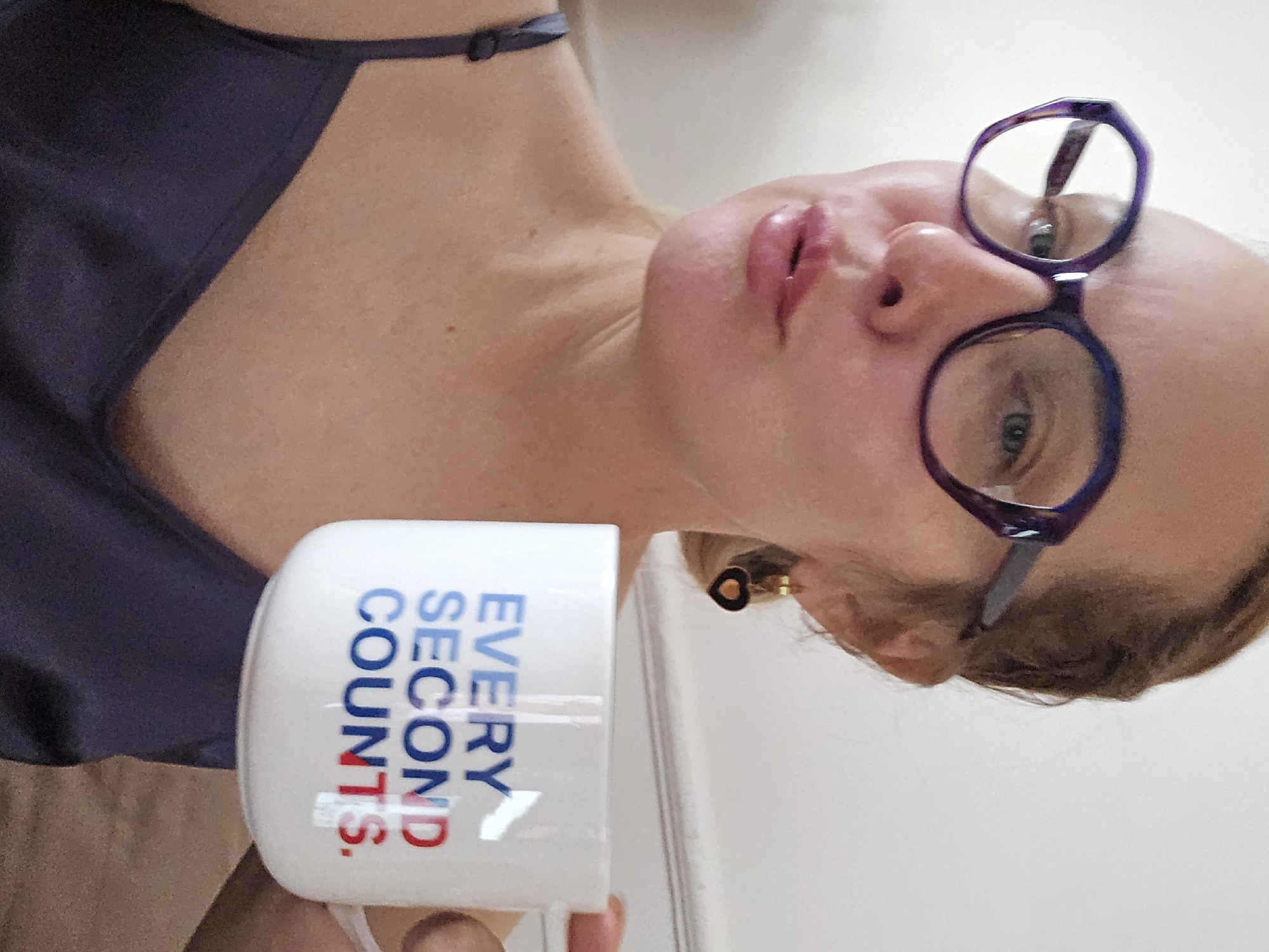 A woman wearing glasses and a dark-colored top is holding a white mug with the text "EVERY SECOND COUNTS." Her face is visible, and she appears to be looking at the camera. She is wearing a small gold earring. The background is a plain white wall. The image is rotated to the left. There is no information to suggest the location of the image is in Sochi, Russia.