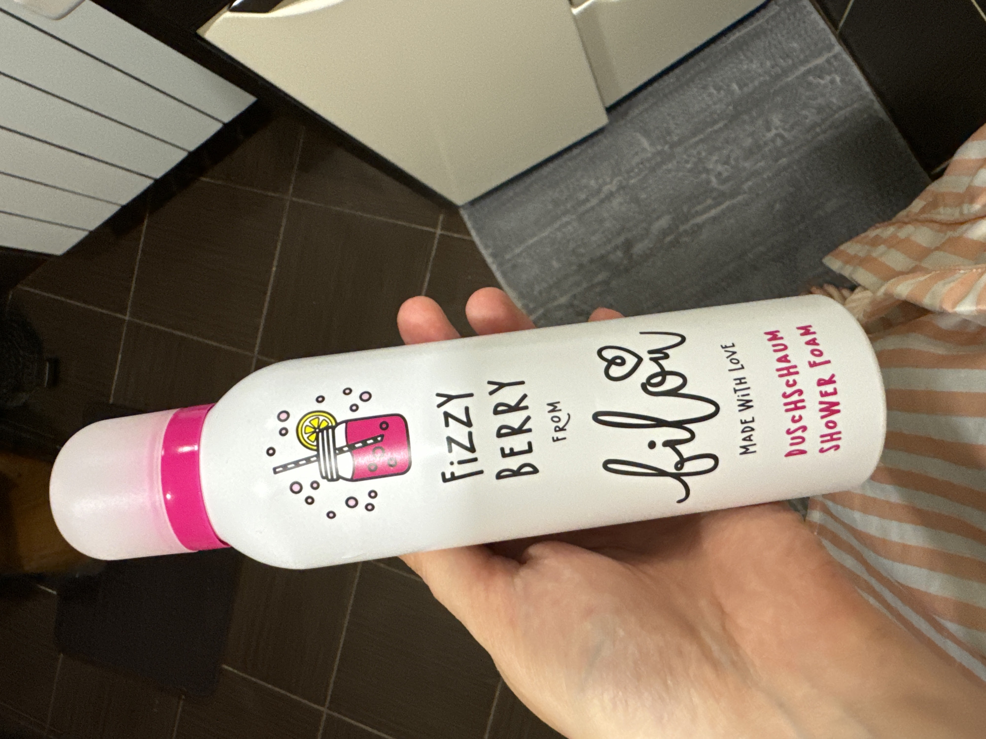 A person's hand is holding a white bottle of "Fizzy Berry Shower Foam" by "bilou". The bottle has a pink cap and a drawing of a jar with a straw and a lemon slice. The background shows a tiled floor and a partially visible white surface. The scene is indoors and appears to be in a bathroom.