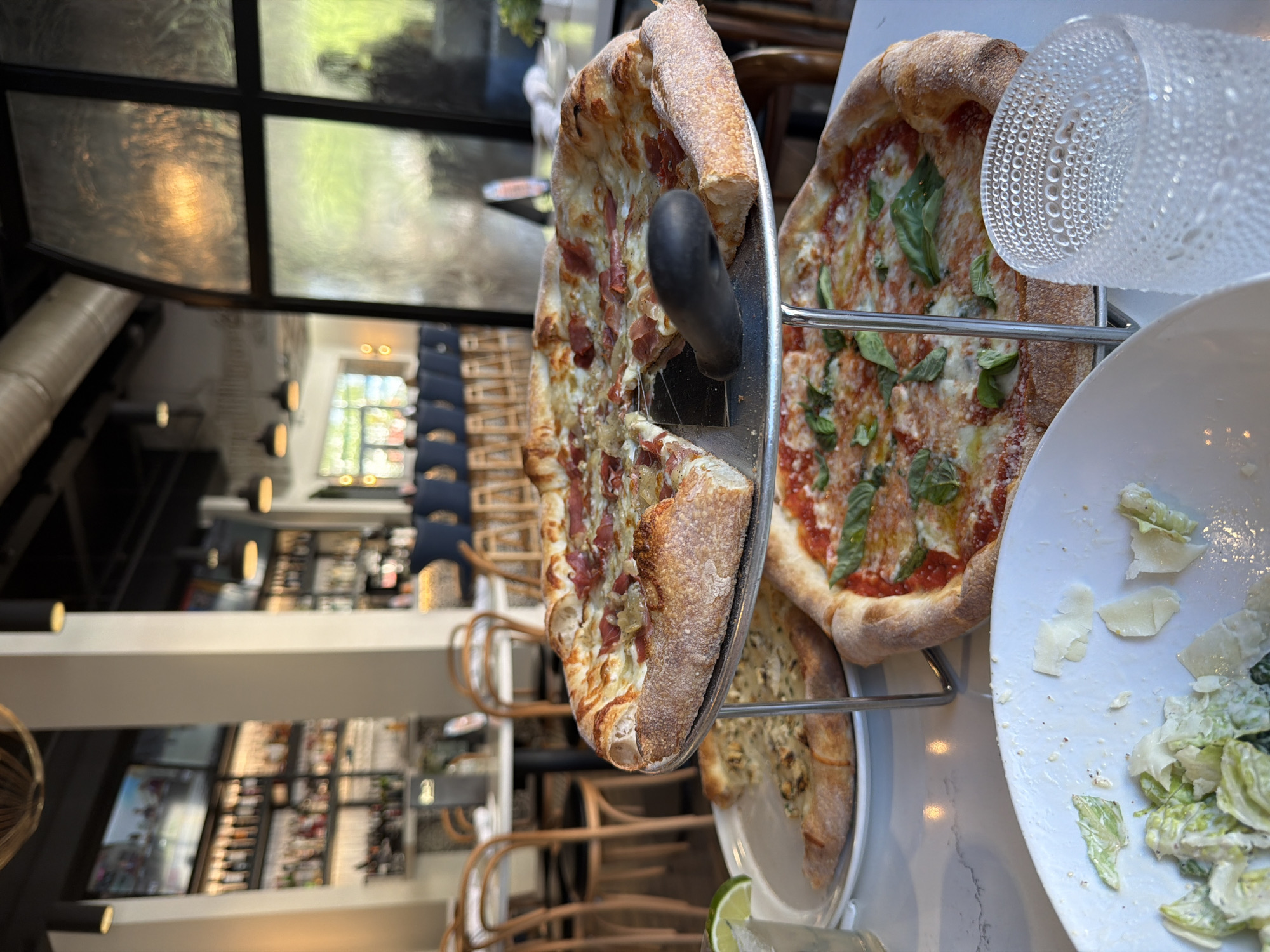 The image shows a food scene in what appears to be a restaurant. There are three pizzas displayed on a tiered metal stand. One pizza features a tomato-based sauce with basil leaves. Another pizza has a white sauce base with cured meat. The third pizza is topped with what appears to be mushrooms. To the side of the pizzas is a partially eaten salad on a white plate. A water glass and lime wedge are also visible. The background indicates an interior restaurant setting. There are chairs, tables, bar-style furniture and stocked shelves of alcoholic beverages. Based on the visible environment, it can be inferred that the photo was taken inside an establishment offering Italian cuisine. It is unclear if this photograph was taken in the city of Myrtle Beach, United States of America, and there are no people visible.