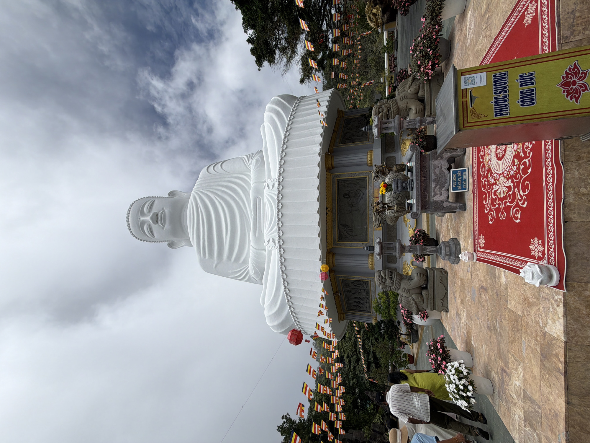 This image shows a large, white Buddha statue sitting on a pedestal at a temple in Đà Nẵng, Vietnam. The statue is very tall and occupies most of the frame, with its head reaching towards the cloudy sky. The Buddha is depicted in a seated meditation pose. In front of the pedestal, there are two stone lion statues, a red carpet with intricate patterns, and several potted plants.
The area around the statue is decorated with strings of colorful flags, predominantly yellow, red, and blue, strung across wires. There are also red lanterns hanging from some of the wires. In the foreground, several people are visible. Two individuals are walking away from the camera towards the statue. One is wearing a striped shirt and dark pants, and the other is wearing a yellow dress. Another person in a blue shirt is partially visible on the left.
A yellow donation box with Vietnamese text and a lotus flower design is positioned on the right side of the image. The ground is paved with large, textured tiles. In the background, lush green trees and vegetation can be seen, suggesting the temple is located on a hillside or in a mountainous area. The overall scene suggests a serene and spiritual atmosphere. The specific location in Đà Nẵng is likely the Lĩnh Ứng Pagoda on Sơn Trà Peninsula, which is famous for its large white Buddha statue.