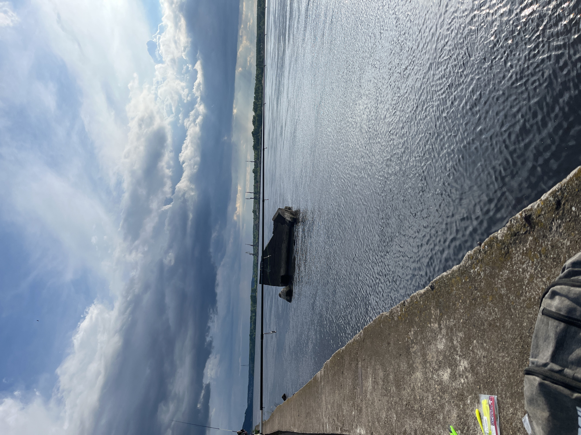 The image shows a water scene, possibly a river or lake, taken from a concrete structure, likely a pier or embankment. The water surface is rippled, reflecting the sky. A large, dark, partially submerged concrete or stone structure is visible in the water. In the distance, across the water, is a strip of land with trees and possibly buildings or structures that appear industrial.
The sky is mostly cloudy, with areas of bright sunlight breaking through.
A fishing rod can be seen in the bottom left of the frame. A backpack and fishing gear are placed on the concrete structure in the lower right corner.
Based on the prompt referencing Chaikovsky, Russia, the water feature is most likely the Kama River, which flows through the city. The structures across the river could be related to industrial activities associated with the city. The setting suggests recreational fishing activity.
