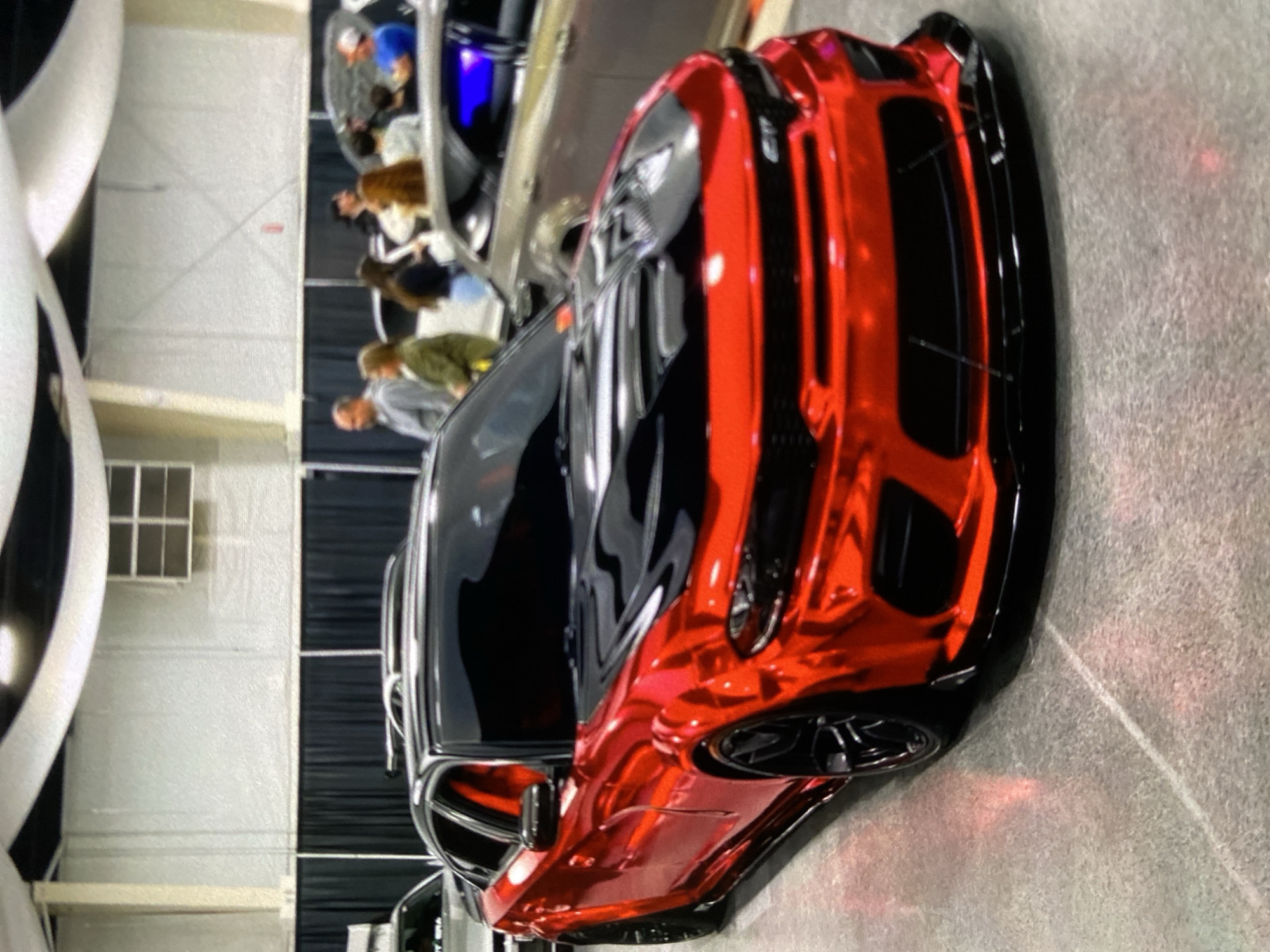 The image shows a red and black car on display at what appears to be an indoor exhibition hall. A crowd of people of varying ages stands in the background, some of whom are near other cars. A window is visible in the background on the left side. Based on the prompt, the image is from Lhokseumawe, Indonesia, but there is no visual information in the image that confirms this.