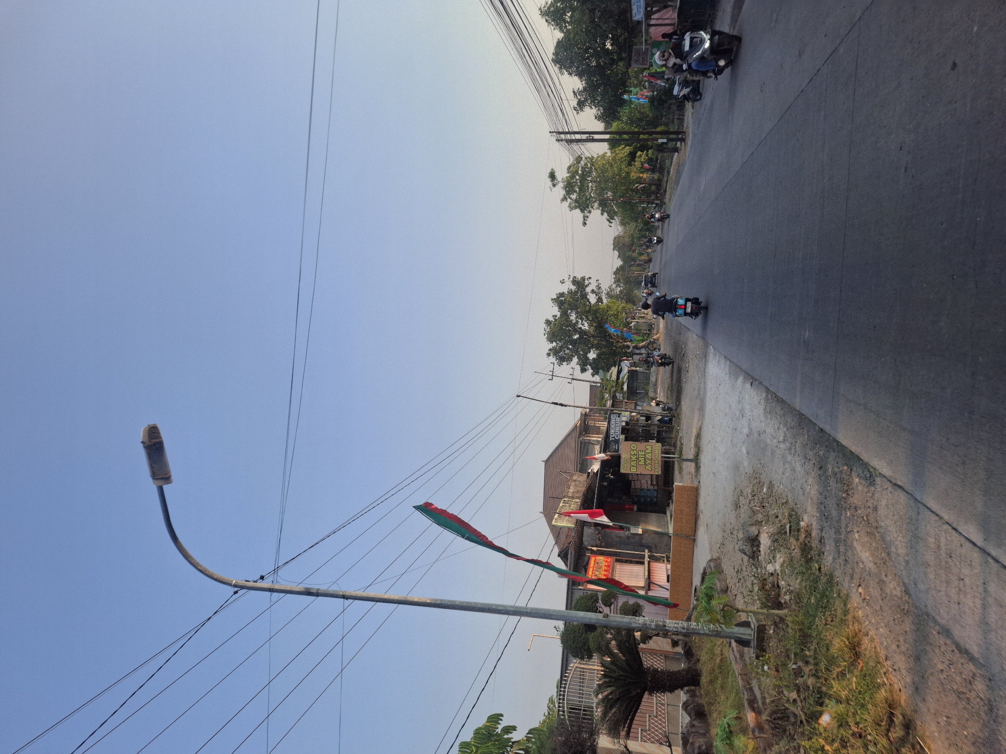 This image depicts a street scene in Banjarmadu, Indonesia, during what appears to be late afternoon. The perspective is from a moving vehicle, looking down a paved road lined with trees and small businesses on the left side.

The sky is a clear, pale blue, with the sun casting long shadows across the road and the vegetation. A prominent streetlamp with a curved pole stands on the left side of the road, adorned with two colorful flags – one green and red, and another red and white Indonesian flag. Below the streetlamp, there are various stalls or shops with some merchandise visible, and signs in Indonesian. One sign reads "BAKSO ME AYAM," indicating a food stall.

Several motorcycles are present on the road, with individuals riding them. One motorcycle in the foreground on the right has a rider wearing a helmet and blue jeans. Further down the road, more motorcycles and a few cars are seen in motion. The roadside on the left is a mix of dry grass, some plants, and a decorative concrete barrier. The overall environment suggests a relatively calm, everyday street scene in a semi-urban or rural area.