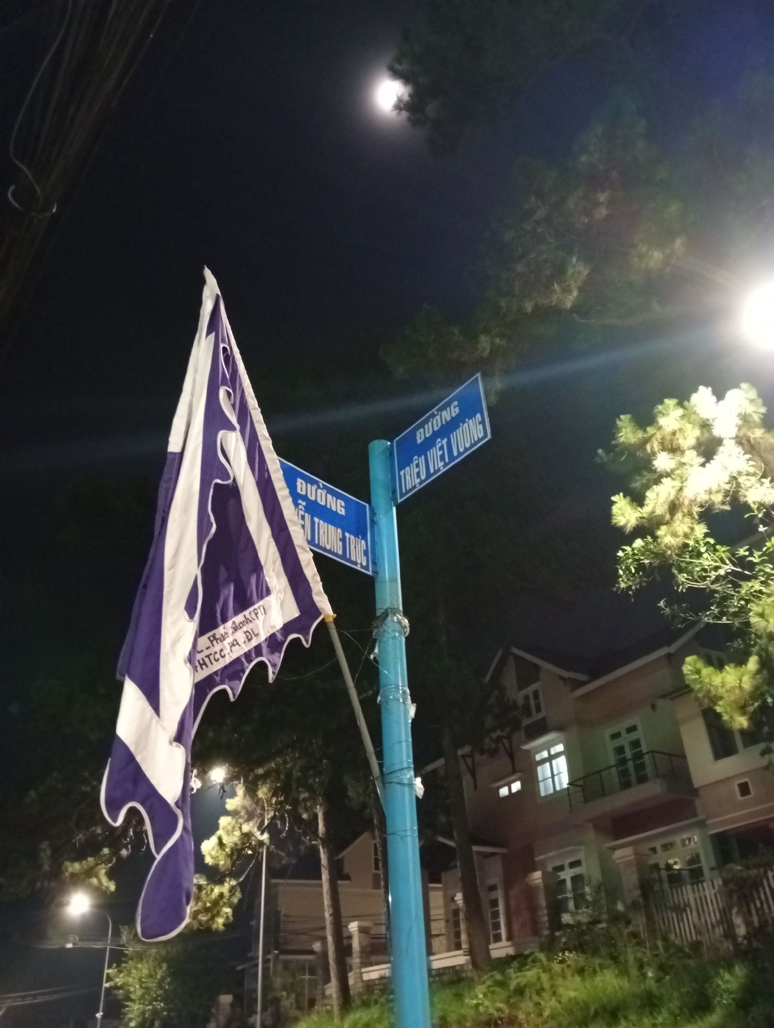 The image shows a street scene at night in Da Lat, Vietnam. A blue street signpost indicates the intersection of "ĐƯỜNG TRIỆU VIỆT VƯƠNG" and "ĐƯỜNG HIẾN TRUNG TRỰC". Attached to the signpost is a flag with purple and white stripes, bearing text that includes "Phước Thanh (PT) HTCC PA DL". A brightly lit street lamp illuminates the area around the signpost and the house in the background, where windows are visible on two floors. Trees are visible to the side, and a full moon is visible in the sky. Electrical wires are strung overhead.