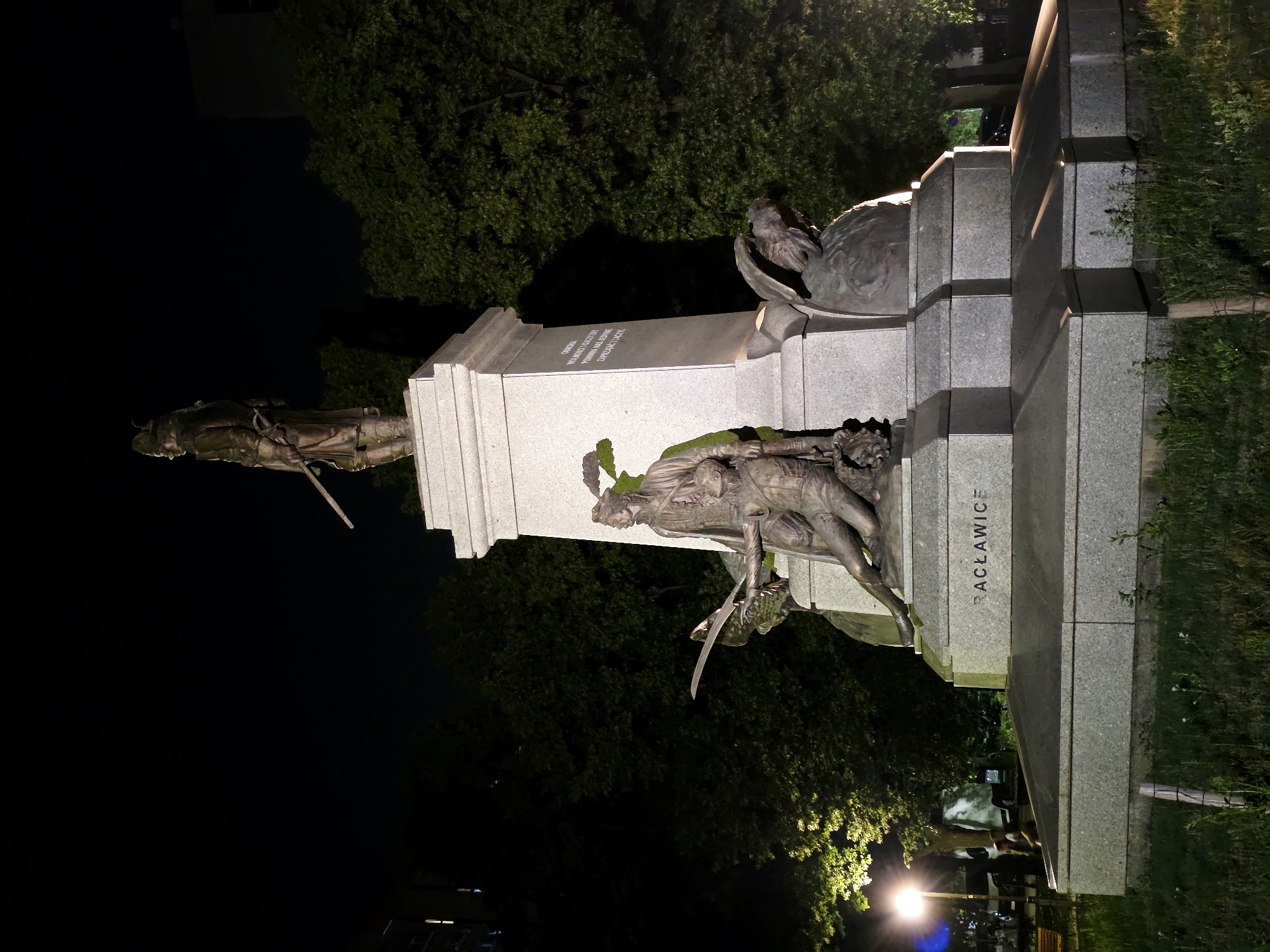 The Racławice Panorama is a monumental circular painting, 114 meters long and 15 meters high, depicting the Battle of Racławice. It is housed in a purpose-built rotunda in Wrocław, Poland. The image provided shows a monument at night, illuminated by artificial lights. The monument features bronze sculptures of soldiers and an eagle, set against a stone pedestal. Text on the pedestal reads "RACŁAWICE". Trees and a street lamp are visible in the background. The scene is set outdoors, likely in a public space such as a park or square. The monument appears to commemorate a historical event, likely the Battle of Racławice.