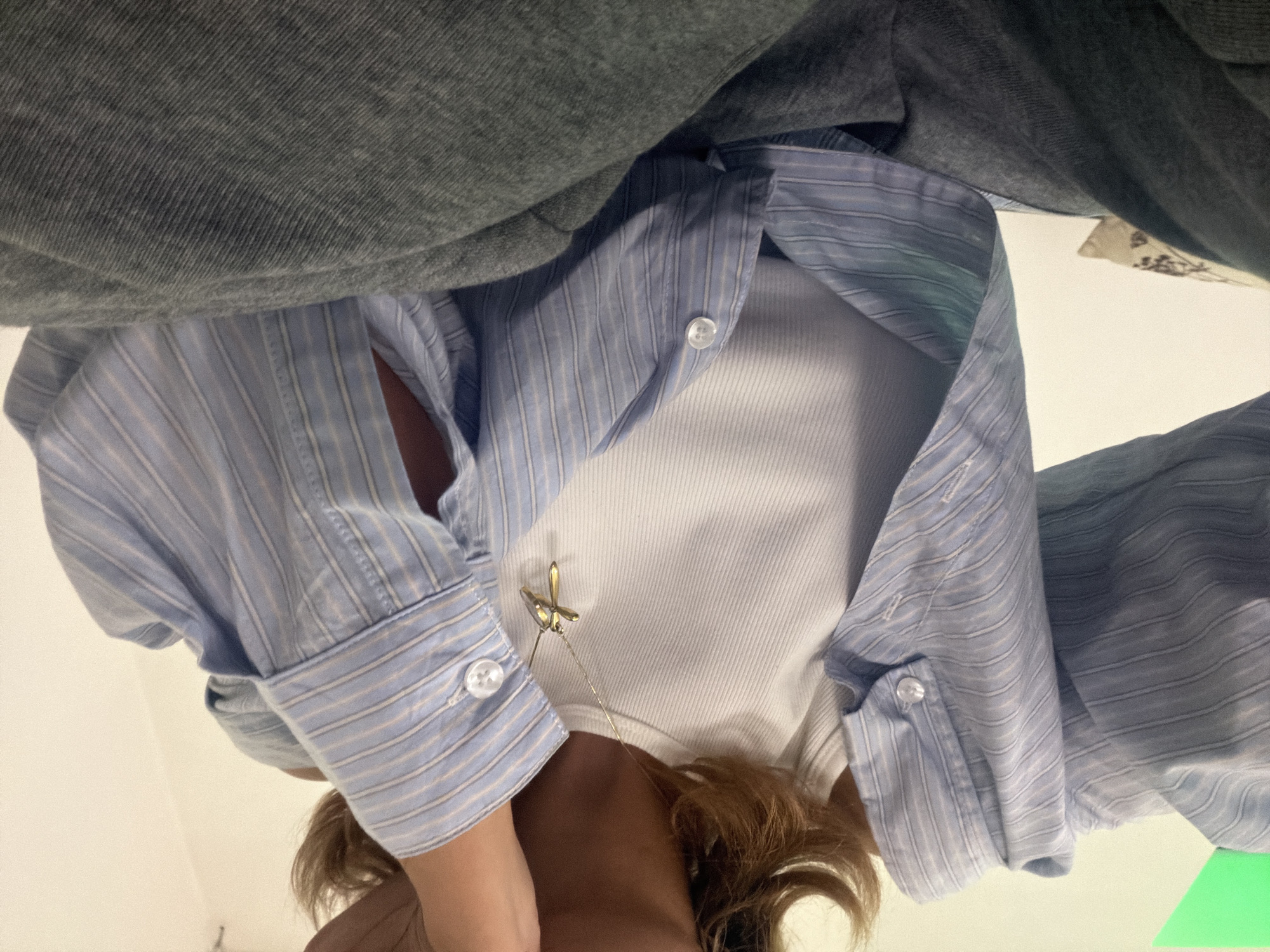 The image presents a view looking down on a person wearing a white ribbed tank top, a light blue and white striped collared shirt, and a grey sweatshirt. A delicate gold necklace with a pendant resembling a dragonfly or stylized flower is visible. The angle of the photograph implies it was taken from a high vantage point looking straight down. The location in Yerevan, Armenia is not apparent from the available visual information.
