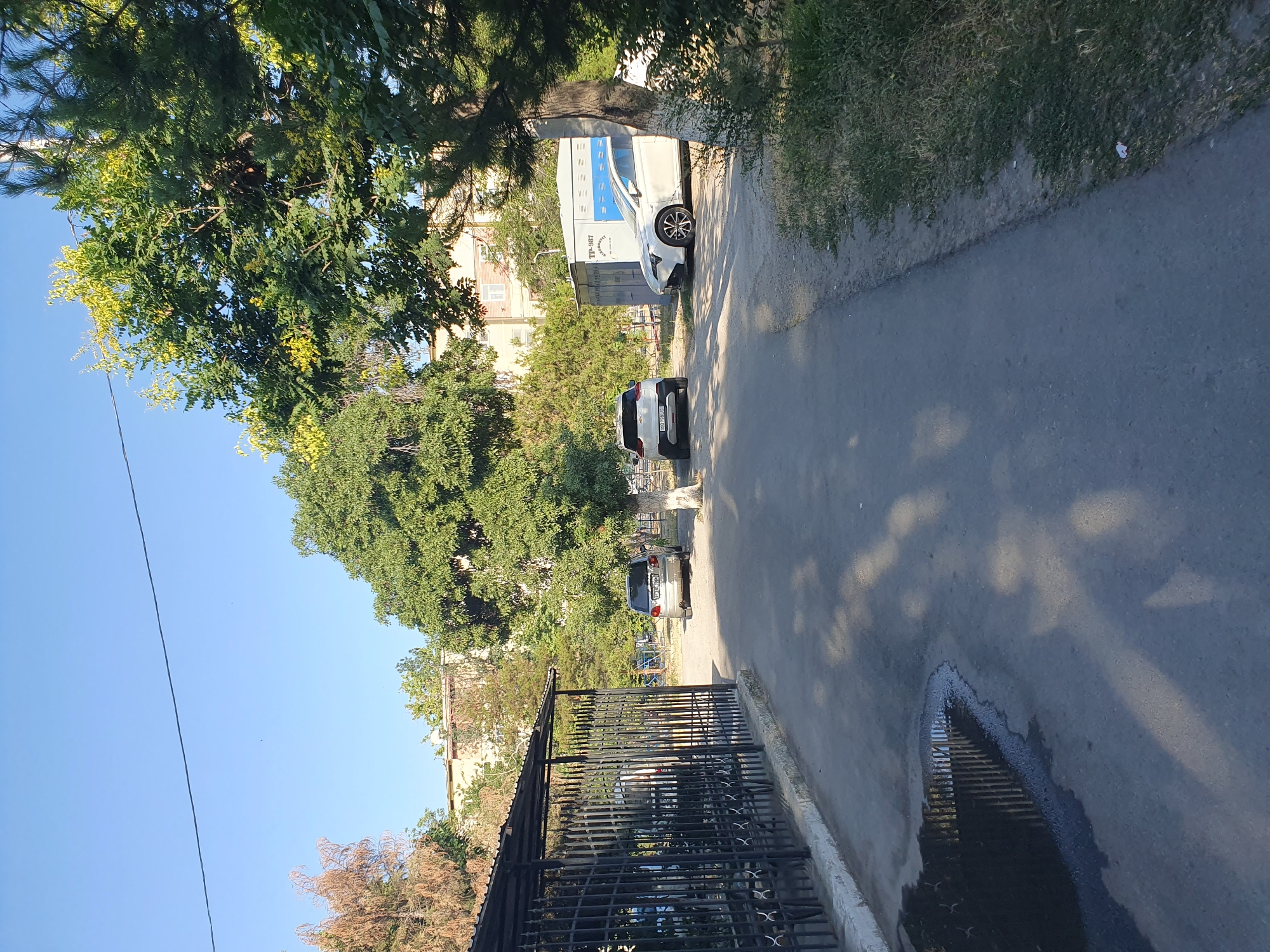This is a daylight, eye-level, medium shot of a street scene in Tashkent, Uzbekistan. The scene is characterized by a paved road with some shadows cast by trees and a fence. To the left of the road, there is a black metal fence with a roof overhead, creating a covered area. Beyond the fence, some parked vehicles are visible. The road itself has a large puddle of water reflecting the fence. In the middle ground, there are three cars parked: a silver SUV, a white SUV, and a white sedan. Behind the cars, there is a light-colored building with windows and trees surrounding it. The sky is clear and blue. The overall impression is of an urban environment with residential buildings and greenery.
