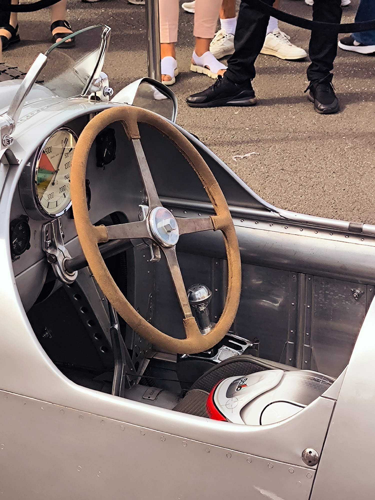 The image is a close-up shot of the interior of a vintage silver race car. The focus is on the steering wheel, dashboard, and a racing helmet. The steering wheel is tan-colored and appears to be made of suede. The dashboard features a large, circular speedometer with a red, green, and yellow dial. To the left of the speedometer is a smaller, black dial. The car's interior is minimalist, with exposed metal paneling and rivets. A white and red racing helmet with black accents rests on the car's seat. In the background, the legs of several people are visible, suggesting an outdoor event. The ground is paved with asphalt. The scene does not contain any specific visual cues to identify the location as Riga, Latvia.