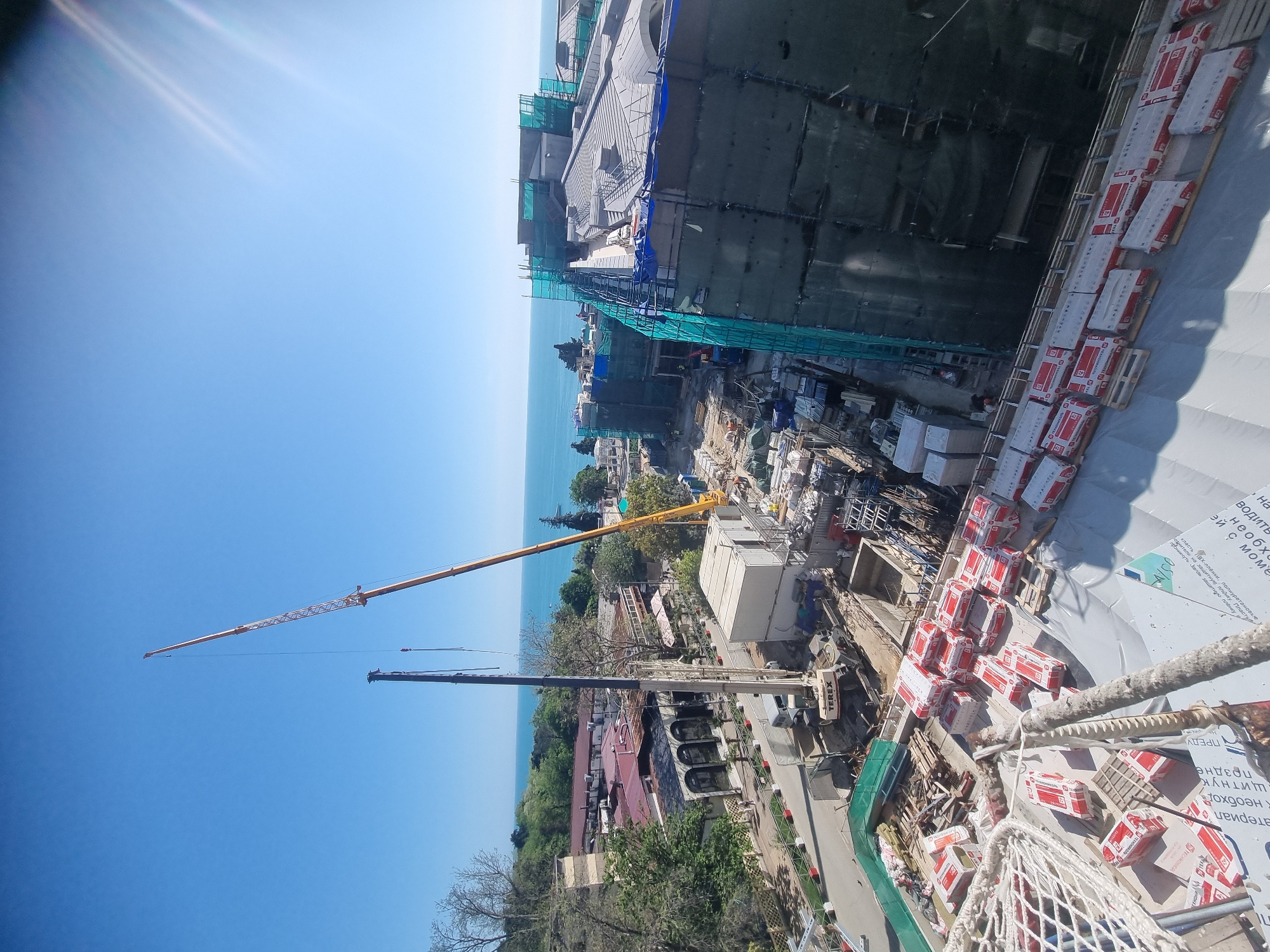 The image shows a construction site in Sochi, Russia, viewed from a high vantage point. The site includes a large building under construction, scaffolding, and building materials such as stacks of red and white packaged items. Construction cranes are visible in the scene, one with a long yellow boom and another with a dark boom. The Black Sea stretches out in the background. The foreground shows parts of the structure the photograph was taken from, including ropes and packaging of building materials.