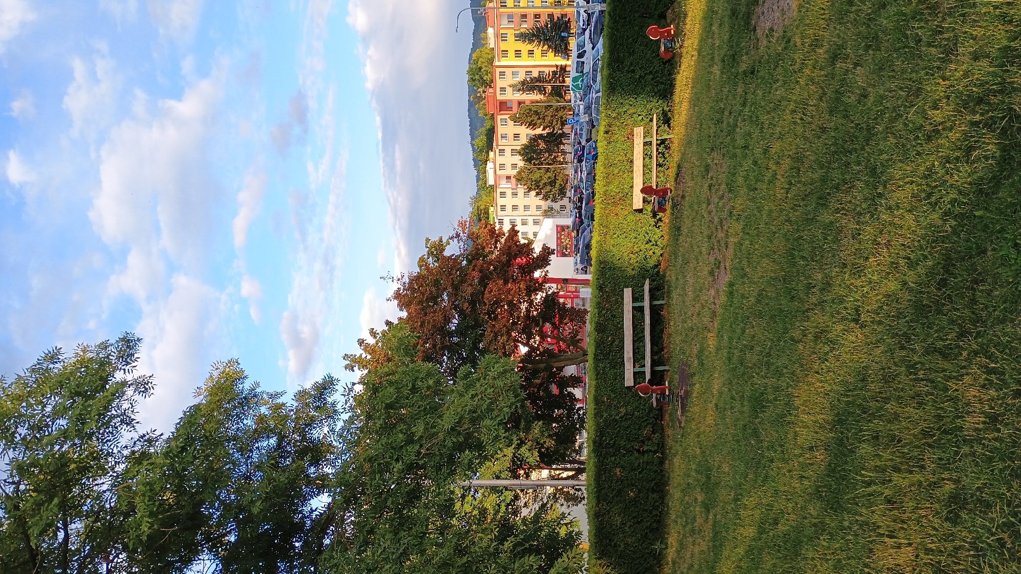 The image depicts a residential area in Sokolov, Czechia. In the foreground, a patch of green grass is illuminated by the late afternoon sun, casting long shadows. Two park benches are visible, one on the left and another on the right, both made of wood and metal. A red, spring-mounted toy, possibly a seesaw or a bouncing toy, is situated in front of the right bench. Behind the benches is a neatly trimmed green hedge, separating the park area from a parking lot. The parking lot is filled with numerous cars, suggesting a densely populated residential block. In the background, several multi-story apartment buildings are visible, painted in various colors, including yellow, orange, and light green. The buildings are bordered by trees, and a few more apartment buildings can be seen in the distance, nestled against a backdrop of rolling hills. The sky is a clear blue with scattered white clouds, indicating fair weather. A street lamp is visible on the right side of the image, near the apartment buildings. The overall scene is a typical view of a residential neighborhood in a European city.