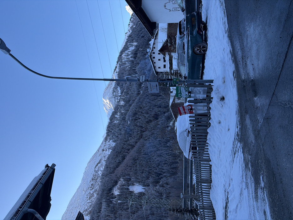 This image captures a snow-covered winter scene in Sankt Jakob am Arlberg, Austria, specifically near a location indicated by a sign for "Gruberhof 1.1". The environment is mountainous, featuring dense, snow-dusted coniferous forests on lower slopes and prominent snow-capped peaks in the background, some displaying visible terracing or avalanche prevention structures. The sky is clear and bright blue.
In the immediate foreground, the ground consists of a wet, dark asphalt surface, likely icy, bordered by drifts of packed snow. To the left, a portion of a dark building eave with several icicles is visible. A snow-covered wooden picket fence extends across the mid-ground. Behind this fence, a dark-colored vehicle is parked, almost entirely covered by a thick layer of snow. Adjacent to it, a red sign with white text reads "Kronen Zeitung".
Centrally positioned is a tall, slender grey streetlamp. Attached to its pole are multiple directional signs; one brown sign clearly points left towards "Gruberhof 1.1" with an arrow, and a smaller green sign is visible below it. To the right of the streetlamp, a dark teal-colored crossover SUV with an Austrian license plate (partially visible as "0M-FA-9") is parked, also dusted with snow. Several traditional white-walled buildings with dark roofs are interspersed among the trees in the mid-distance. Power lines are visible stretching across the sky. No human figures or overt human activity are present in the scene.
