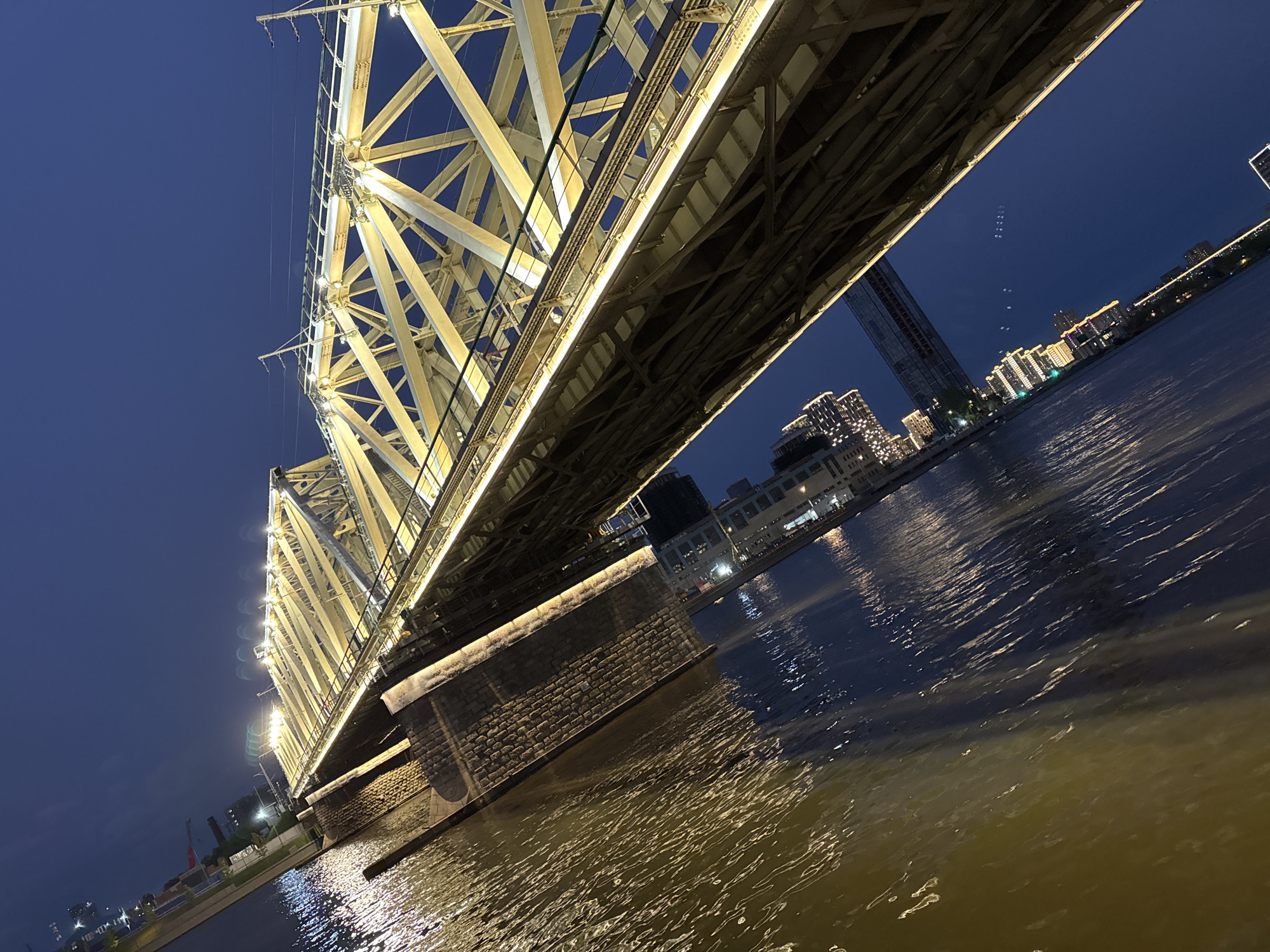 View from below the illuminated Novospassky Bridge in Moscow, Russia, at dusk. The bridge’s steel lattice structure is lit with yellow light. The river is reflecting the lights from the bridge and nearby buildings. In the background are buildings and a tower. The sky is a deep blue. A pier supporting the bridge is made of stone blocks.