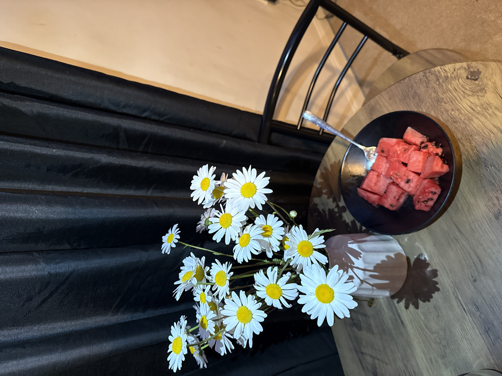 The image shows a still life scene on a wooden table. A vase of white daisies is positioned next to a black plate containing cubed watermelon, with a fork resting on the plate. The table is next to a black metal bed frame and a black draped background. The location is not discernible from the image.