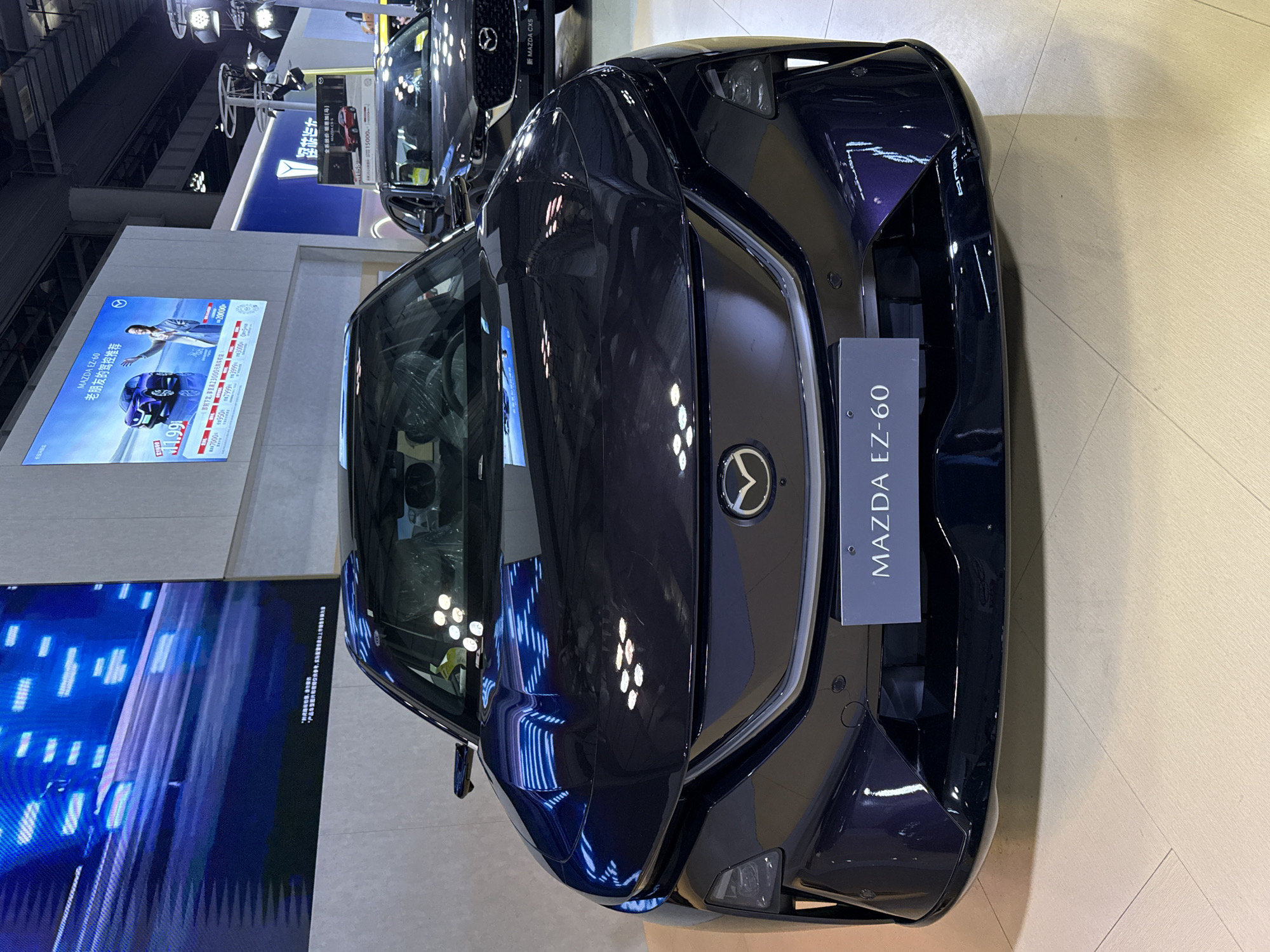 Here is a detailed description of the image:
**General Overview:**
The image showcases the front view of a dark blue Mazda EZ-60 car displayed at a promotional event or an automotive exhibition. The scene is set indoors, likely within a convention or exhibition hall.
**Specific Elements:**
* **Car:** The main subject is the Mazda EZ-60. It's positioned to face the camera directly, highlighting its front design. The car is shiny and reflects light in various spots.
* **Signage:** There is a sign attached to the car's front, vertically displaying "MAZDA EZ-60" in a vertical orientation.
* **Display Board:** Behind the car, a large display board or poster is visible. It features a promotional advertisement with text and images. Another board is visible near the upper left corner.
* **Flooring:** The car is placed on a beige or light-colored floor.
* **Surrounding Environment:** The setting is indoors, and the other elements indicate this is an exhibition booth or part of a larger marketing event.
**Location:**
The location is not identifiable in the image; it is only specified in the prompt that the city is Unknown, Unknown.