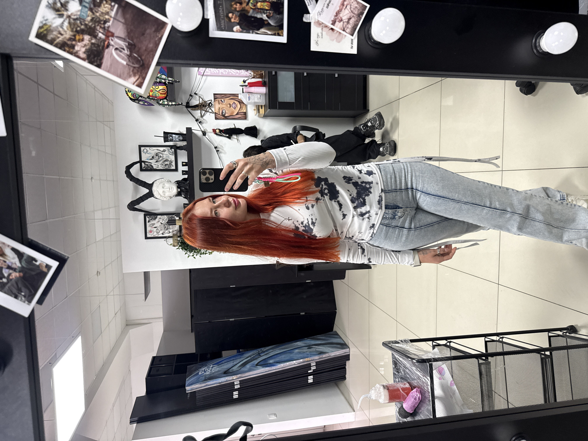 A person with red hair stands facing a mirror, holding a smartphone to take a selfie. They wear light-colored jeans, a white long-sleeved shirt with a dark tie-dye pattern, and white sneakers. A tattoo is visible on their left hand. Reflected in the mirror, a second person lies on the floor. The mirror is part of a vanity with lights attached.
The location is a room with white tiled floors and white walls decorated with framed artwork, photographs, and decorative objects. The room contains black furniture, including cabinets and a rolling cart with art supplies. A stack of artwork is placed on top of a cabinet. The scene is likely within an art studio or salon, but the exact location within Rostov-on-Don, Russia, is impossible to determine with the given information.