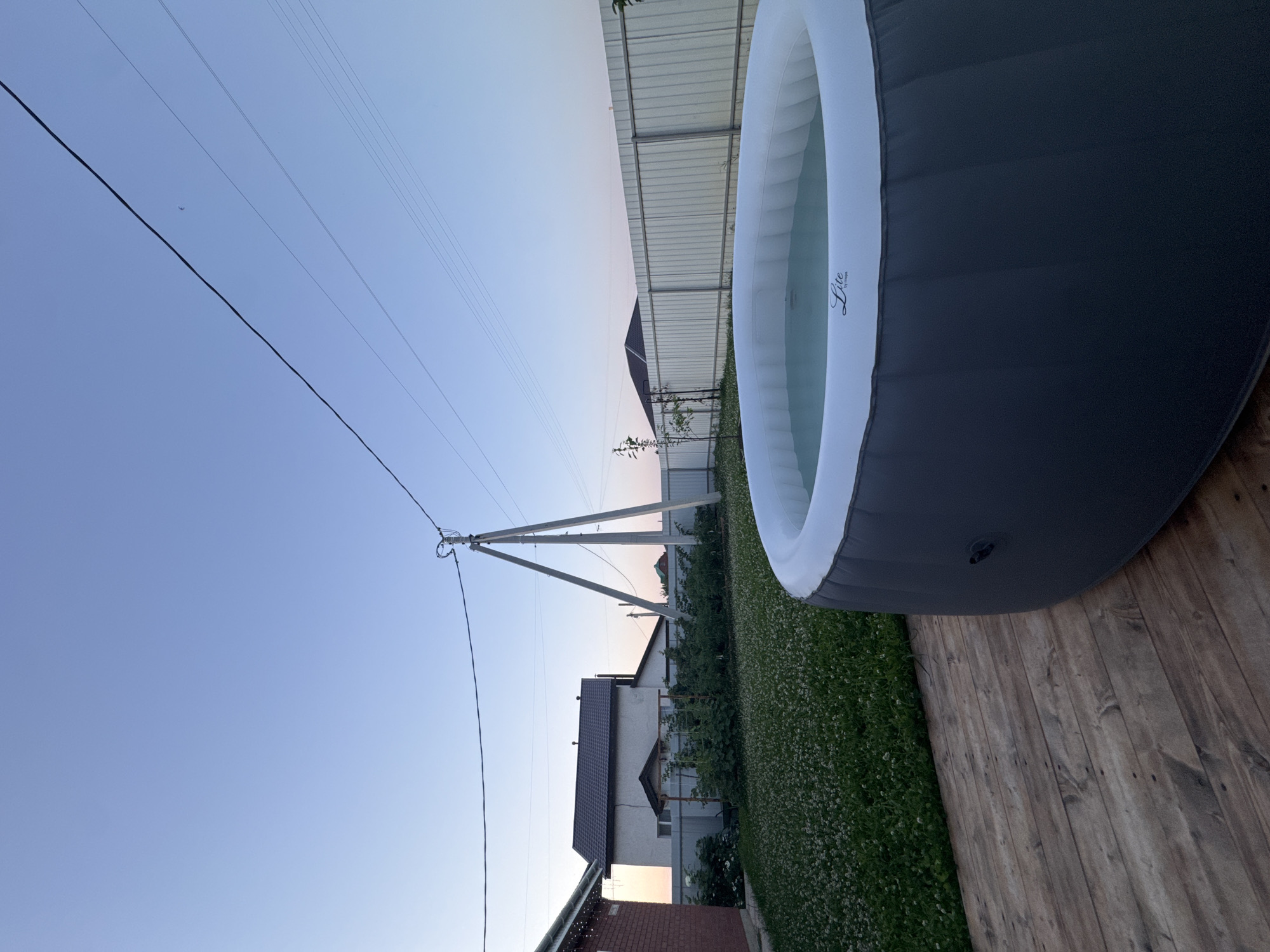 A low-angle, exterior shot captures an inflatable hot tub on a wooden deck. The hot tub is dark gray with a white inflatable rim. A light blue liquid fills the tub, and the brand "Lite by MSpa" is visible on the inner rim. Beyond the deck, a meticulously kept lawn is dotted with white clover flowers. In the background, a light gray metal fence encloses the yard. A house with a dark roof and white siding is partially visible behind the fence, as is a brick house to the left. Utility poles and power lines stretch across the clear, pale blue sky, which suggests it is either dawn or dusk. The overall scene is peaceful and idyllic.