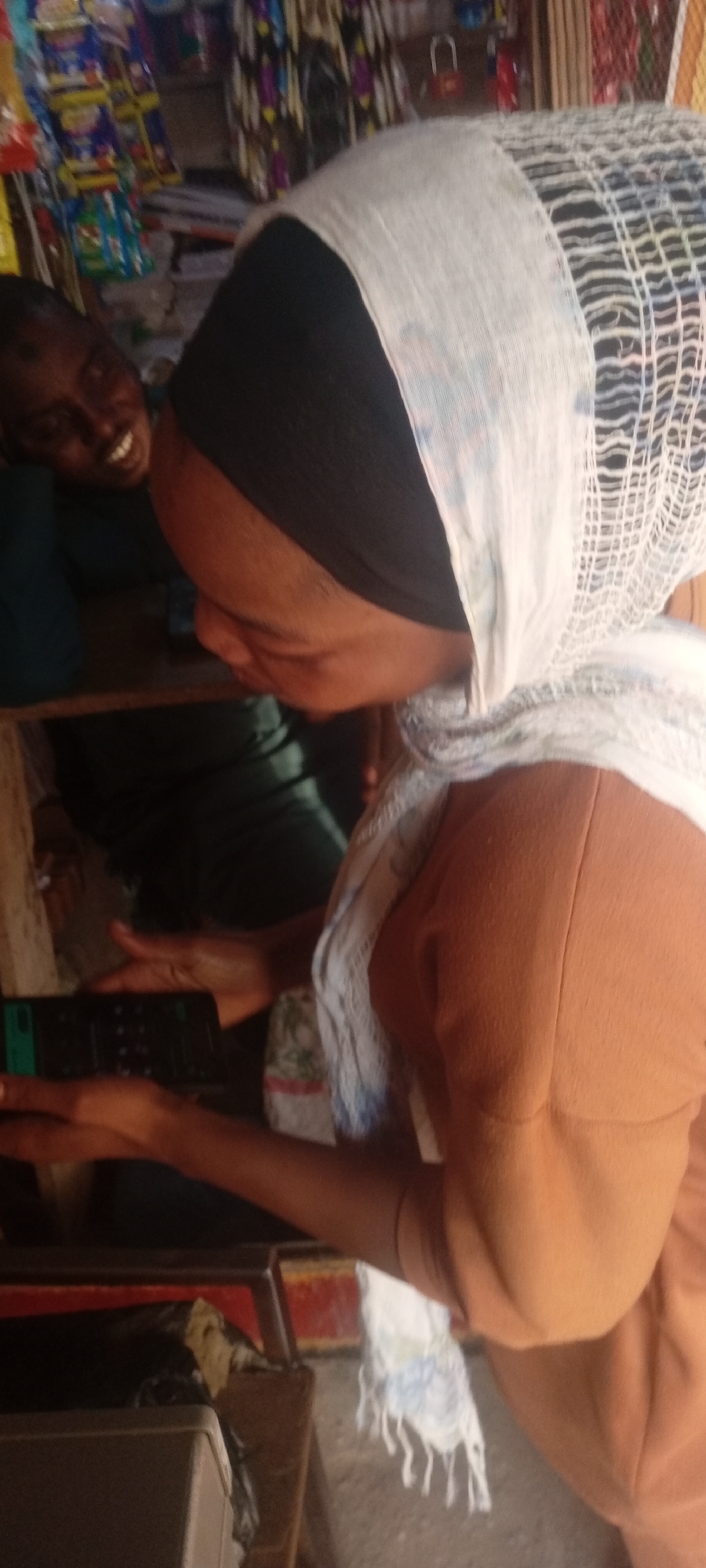 The image shows a woman with a black head covering and a white patterned scarf. She is holding a cell phone in her hands and looking down at it. Behind her, another person is smiling. The setting appears to be a small shop or stall, as indicated by the merchandise in the background. The image was taken in Rijiyar Zaki, Nigeria.