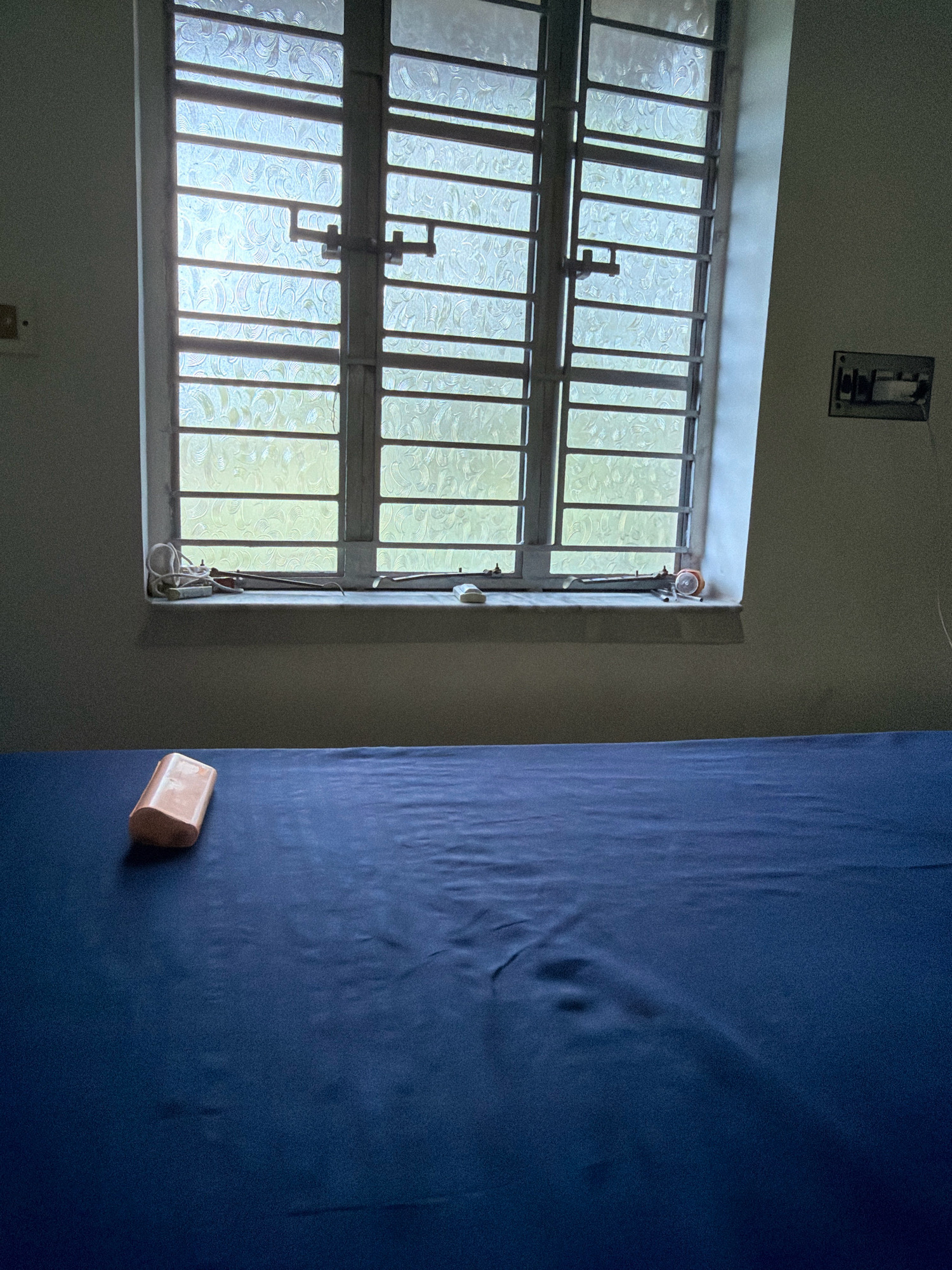 The image depicts a dimly lit room with a large window covered by patterned glass and horizontal bars. On the windowsill, several objects are visible, including what appears to be a small alarm clock, some wires, and a bar of soap. In the foreground, a blue sheet covers a surface, likely a bed, with a light-colored bar of soap resting on it. The overall impression is one of a simple, possibly institutional setting in Kriparampur, India.