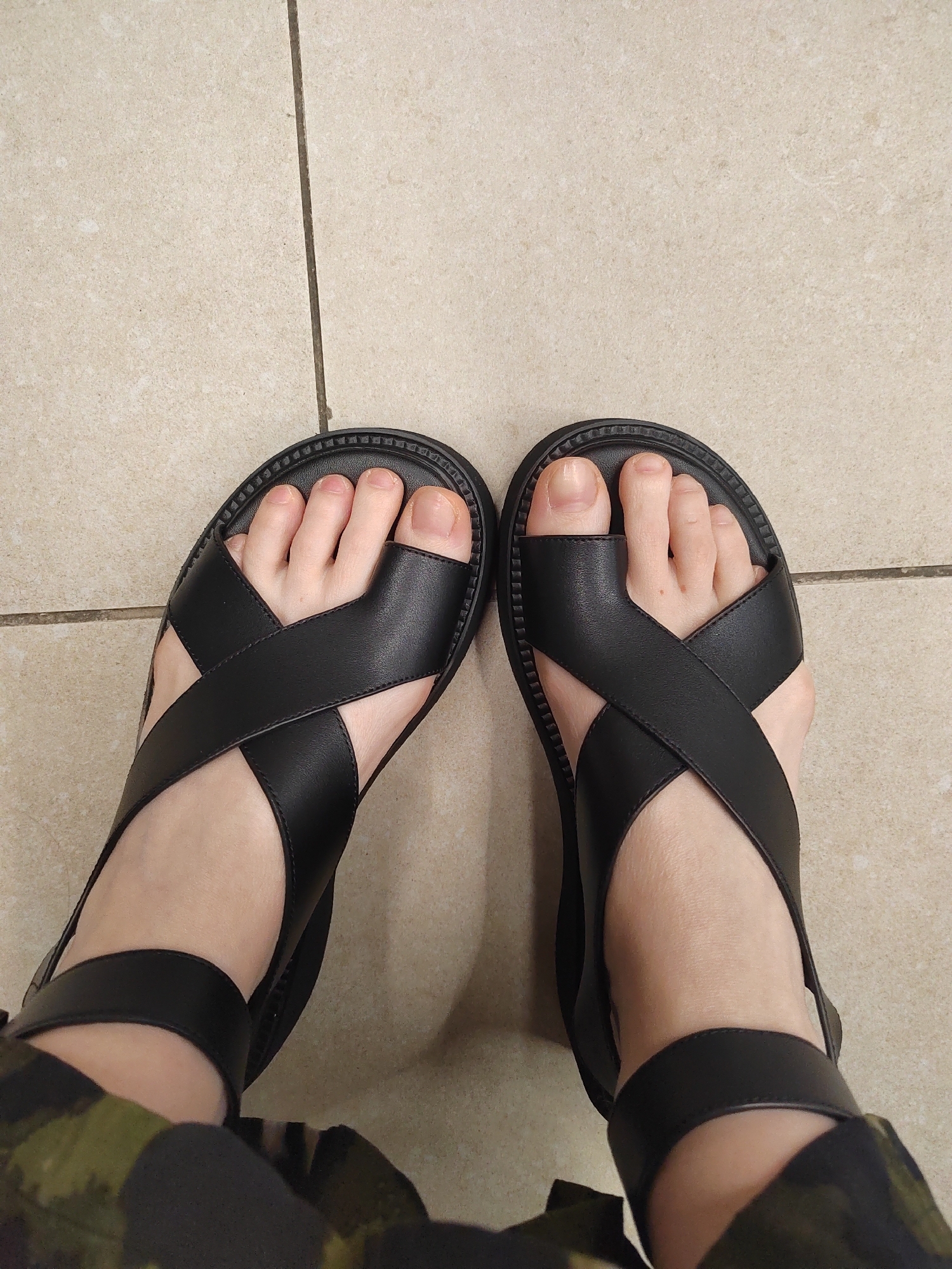 A top-down, medium shot shows a person's feet wearing black cross-strap sandals against a beige tiled floor. The sandals have thick, ridged soles and the straps crisscross over the top of each foot, with one strap encompassing the ankle. The person's toenails are unpolished and their skin is pale. In the foreground, camouflage-patterned fabric is visible, suggesting the lower part of pants or a skirt. The tiled floor appears to be indoors, likely in a residential or public space, with visible grout lines between the tiles. The lighting is even and does not cast strong shadows. There is no indication of the location being in the city of Novogorelovo, Russia.