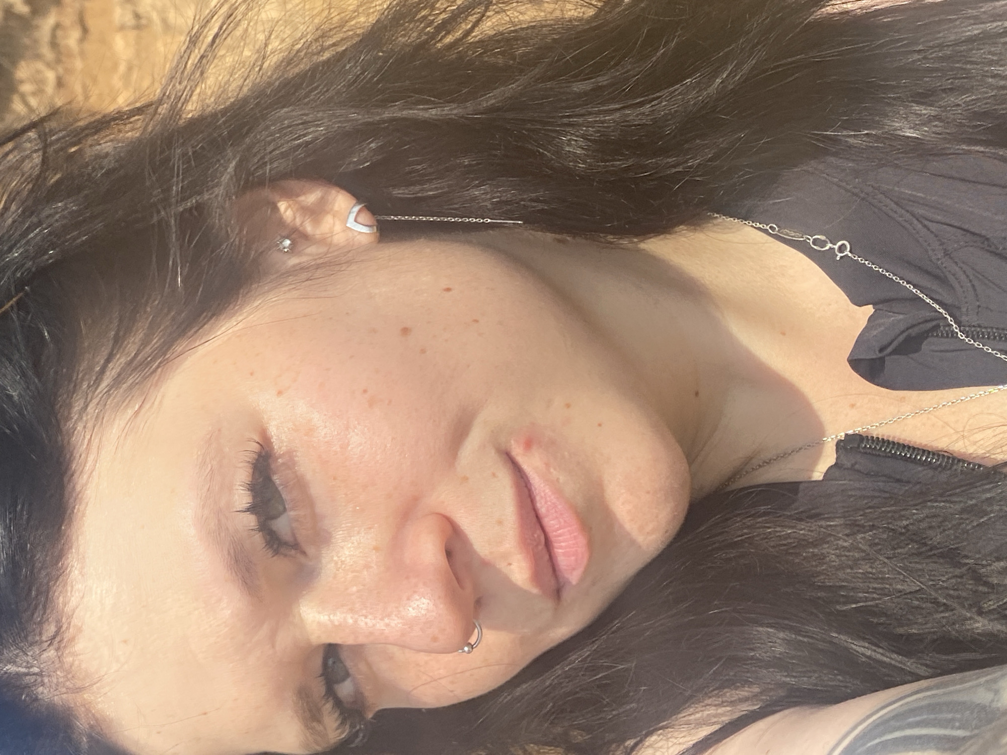 A close-up, eye-level, outdoor, natural light shot features a woman with long, dark hair, facing slightly to the left. Her left eye is visible, with dark eyelashes. She has a silver hoop nose ring and a silver stud in her upper left ear. She is wearing a silver earring with a thin chain that extends down her neck, and a silver necklace with a pendant. She has freckles on her cheeks and a small red blemish on her upper lip. Her hair is dark and long, framing her face. She is wearing a black top with a zipper. The background is out of focus, showing a textured, sandy or rocky surface in warm, natural light, consistent with an outdoor environment in Novyi, Russia.