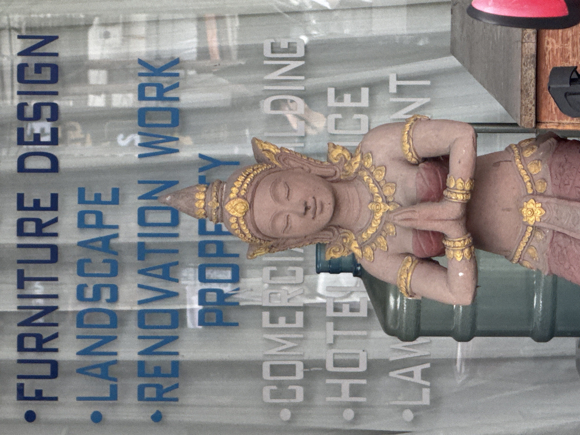 The image shows a storefront in Chalong, Thailand, with text written on the glass. The text, in blue, reads "FURNITURE DESIGN," "LANDSCAPE," "RENOVATION WORK," and "PROPERTY." Below this, in white text, are "COMERCIAL BUILDING," "HOTEL," and "LAW." A statue of a traditional Thai dancer, adorned with gold jewelry and headdress, stands in front of the glass, with hands clasped in a prayer-like gesture. A large green water cooler is visible behind the statue. To the right, a wooden structure with a red object on top is partially visible. The background appears to be a curtain or blinds. The lighting suggests it is daytime.