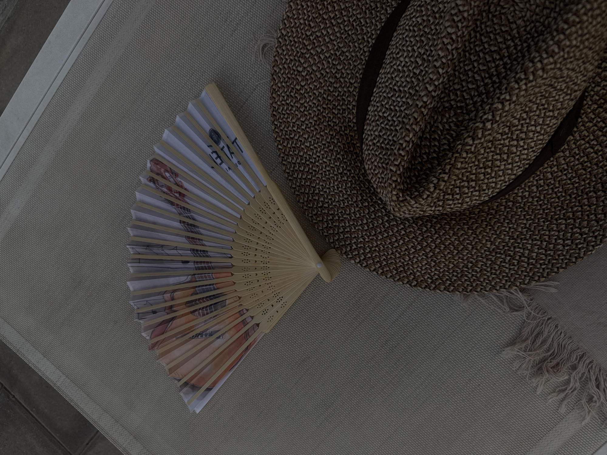 On a textured grey surface, a brown woven hat sits next to a partially open fan. The fan features a printed image. The scene appears to be set outdoors, possibly in a lounge area. There's no indication of the location being in Business Bay, United Arab Emirates. There are no people visible.