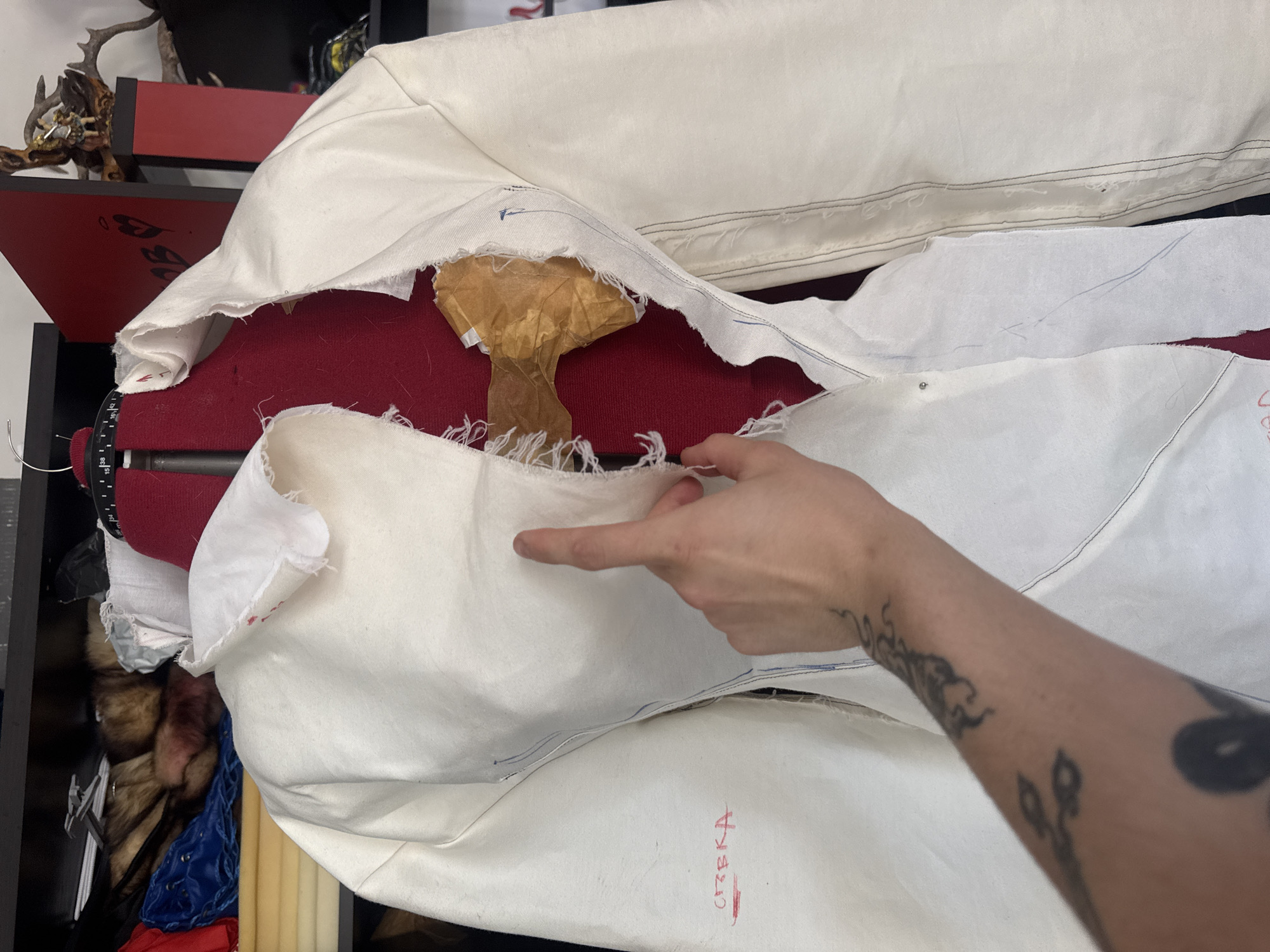 In a cluttered room, possibly a home studio in Sofia, Bulgaria, a person's left arm with visible tattoos is positioned over a partially constructed white garment draped on a red dress form. The garment, possibly a jacket or blouse, is made of unfinished fabric with raw edges and visible stitching. A brown piece of material, likely interfacing or a pattern piece, is taped inside the garment's opening. The background reveals shelves stocked with various items, including fabric scraps, what appear to be fur or faux fur accents, and decorative objects. The lighting is somewhat dim, suggesting an indoor setting.