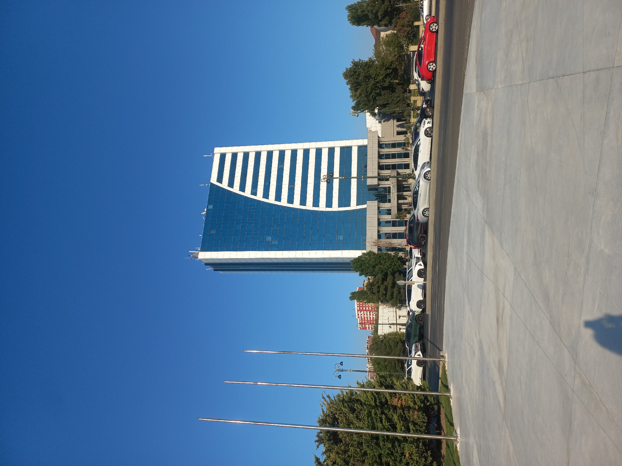 The image shows a modern glass and white-paneled skyscraper in Baku, Azerbaijan, under a clear blue sky. The building has a distinctive curved design on one side, with horizontal white stripes. In front of the skyscraper, several cars are parked along a street. Trees and flagpoles are visible in the foreground. The lower part of the building features a more traditional architectural style with columns. There is a large paved area in the foreground, and the shadow of a person holding a camera is visible on the ground. The overall scene depicts a bright, sunny day in a city environment.
