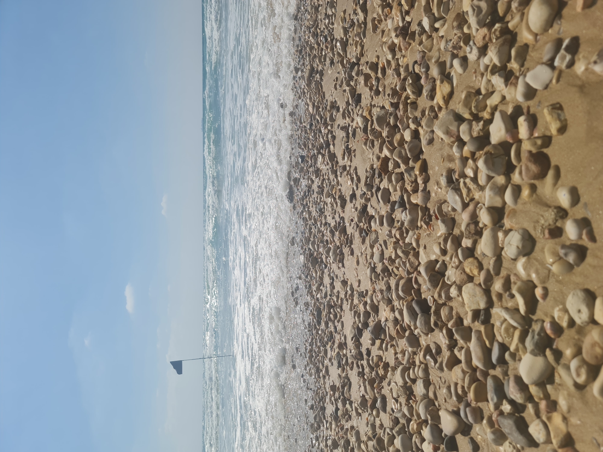 A low-angle, eye-level shot shows a pebbly beach in the foreground, leading to the ocean. The water has moderate waves and white foam. In the distance, a single dark blue flag on a pole is visible in the water, slightly to the left of the frame's center. The sky is clear with a few small white clouds. The scene is brightly lit by natural sunlight.