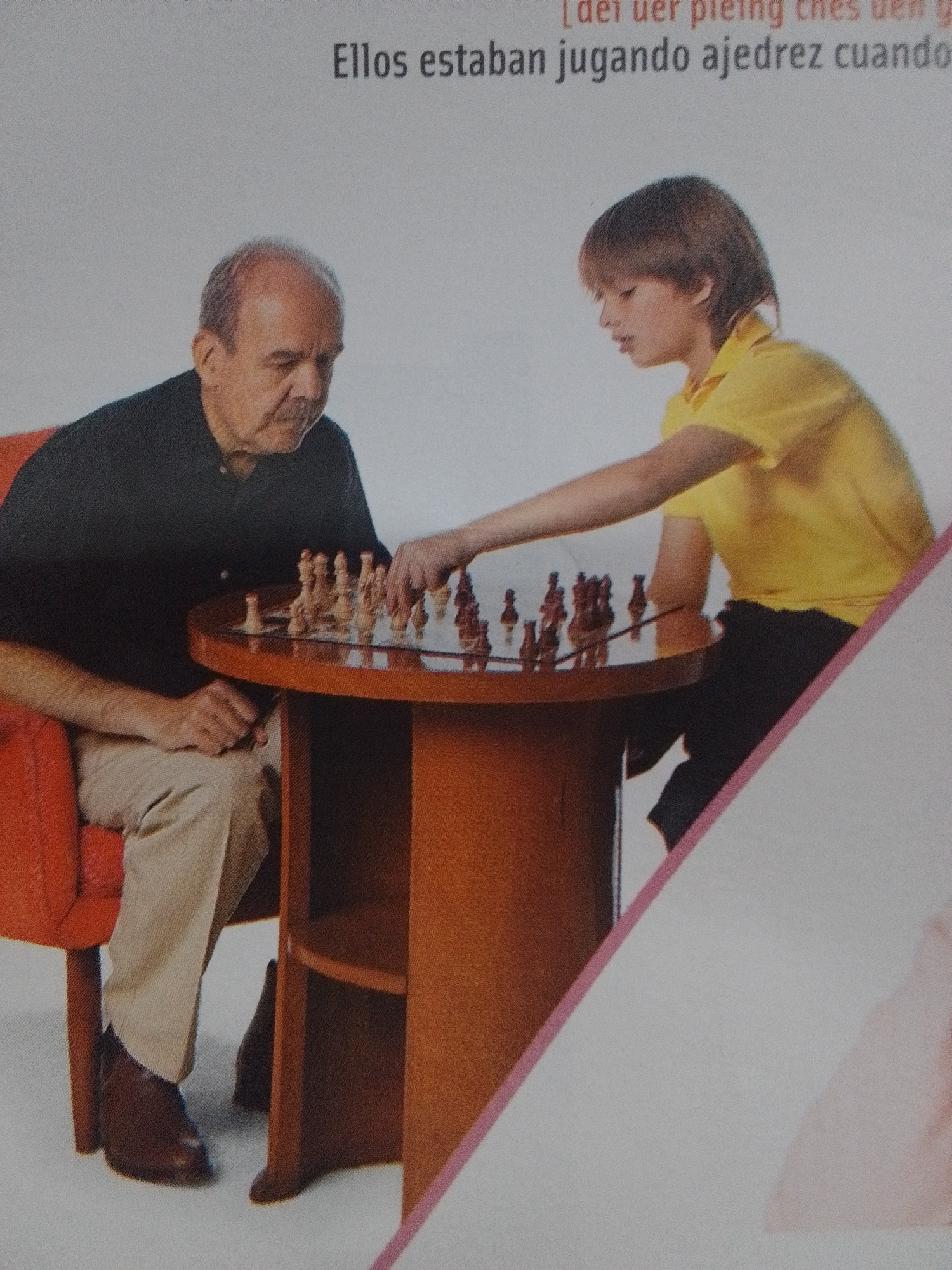 The image shows two individuals playing chess at a round wooden table. On the left, an older man with thinning grey hair and a mustache sits in an orange armchair, wearing a dark blue polo shirt and beige pants. His left hand rests on the table. On the right, a younger person with brown hair, wearing a bright yellow polo shirt and black pants, leans over the table, appearing to be in the middle of making a move with a chess piece. The chess board is set up on the table with wooden pieces. The background is a plain white wall. A partial view of another person, wearing a pink garment, is visible in the lower right corner. The scene is indoors. The image is a photograph, likely taken in La Tagua, Venezuela.