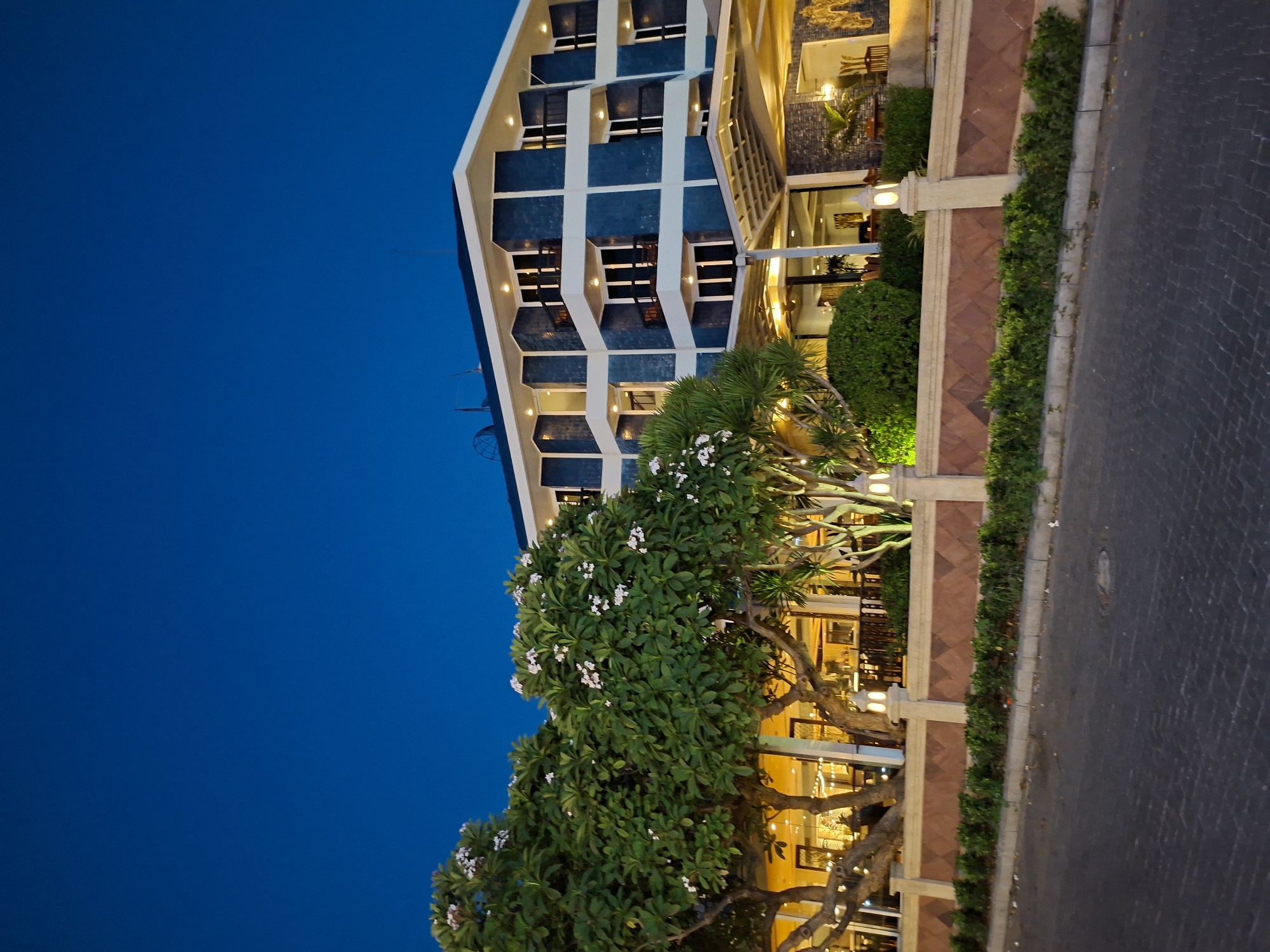 The image depicts a street view at dusk or early evening in Pattaya City, Thailand, featuring a modern building, possibly a hotel, with a blue and white facade. The building has multiple floors with windows and balconies illuminated with warm light.
In the foreground, there is a tree with green foliage and white flowers, its branches partially obscuring the lower section of the building. A low wall runs along the edge of the street, decorated with greenery and interspersed with light fixtures. The street itself is paved with dark-colored bricks or stones. The sky above is a deep blue. No people are visible in the scene.